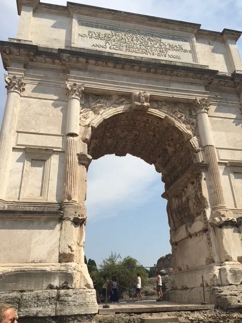 Ancient stone archway with columns and Latin inscriptions, partially damaged and overlooking a few tourists underneath, under a blue sky.