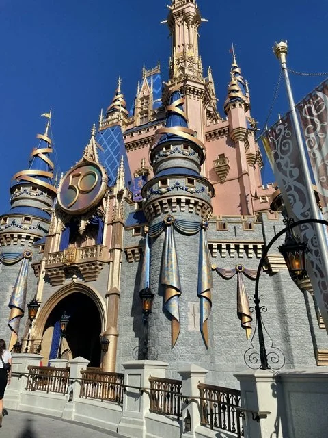Disney castle with pink and blue towers under a clear blue sky, with banners and a clock design on the castle.
