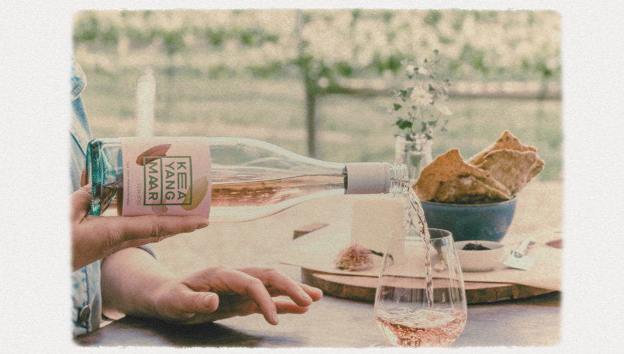 Hand pouring a drink from a pink-labeled bottle beside a glass and a table with crackers in a bright, vineyard-view setting.