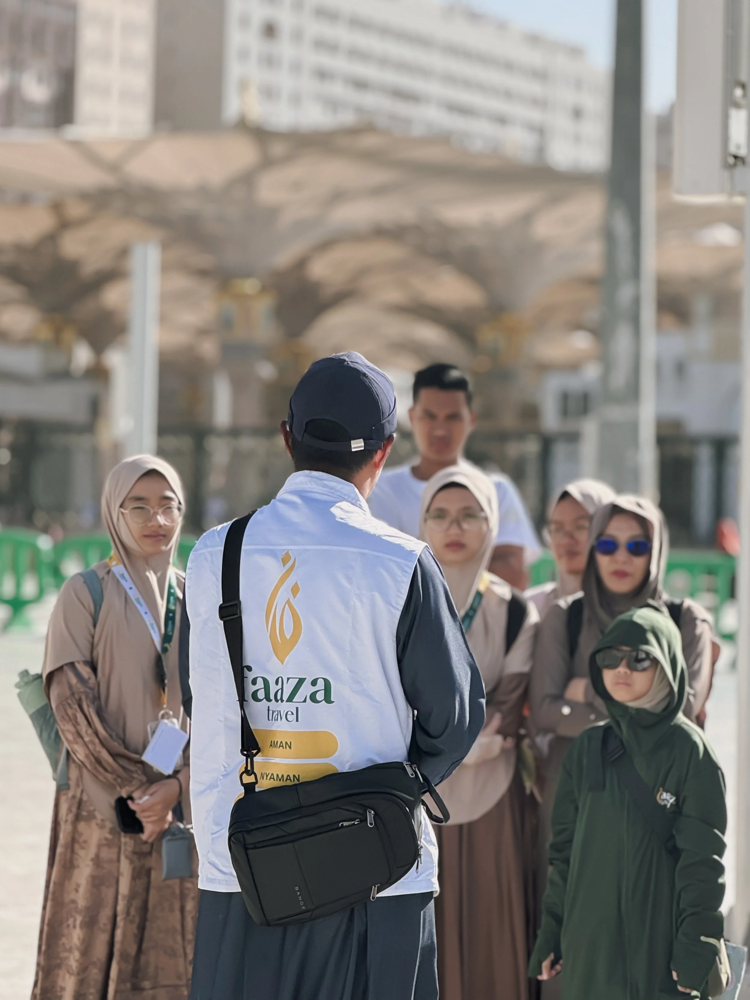 A group of diverse people gathered outdoors, engaging in conversation with a man in the foreground wearing a white vest with an Arabic script and travel-related words on it, and a gray cap. The background includes blurred buildings and urban structur