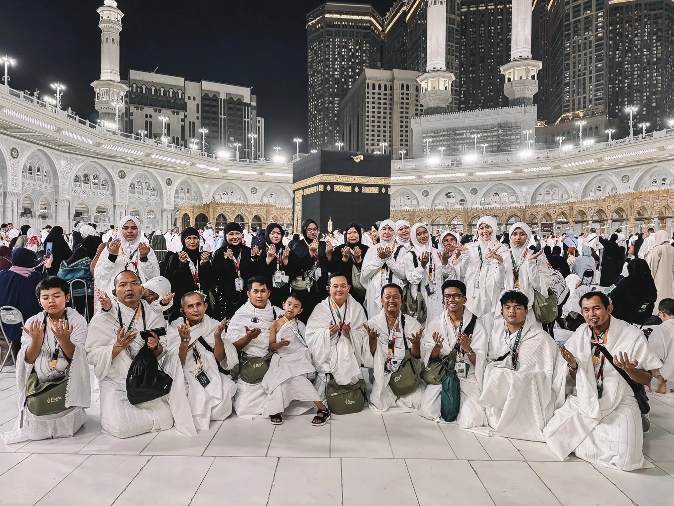 A diverse group of Muslim pilgrims, including men, women, and children, gathered at the Masjid al-Haram in Mecca, Saudi Arabia, near the Kaaba during nighttime with the illuminated mosque and surrounding skyscrapers in the background.