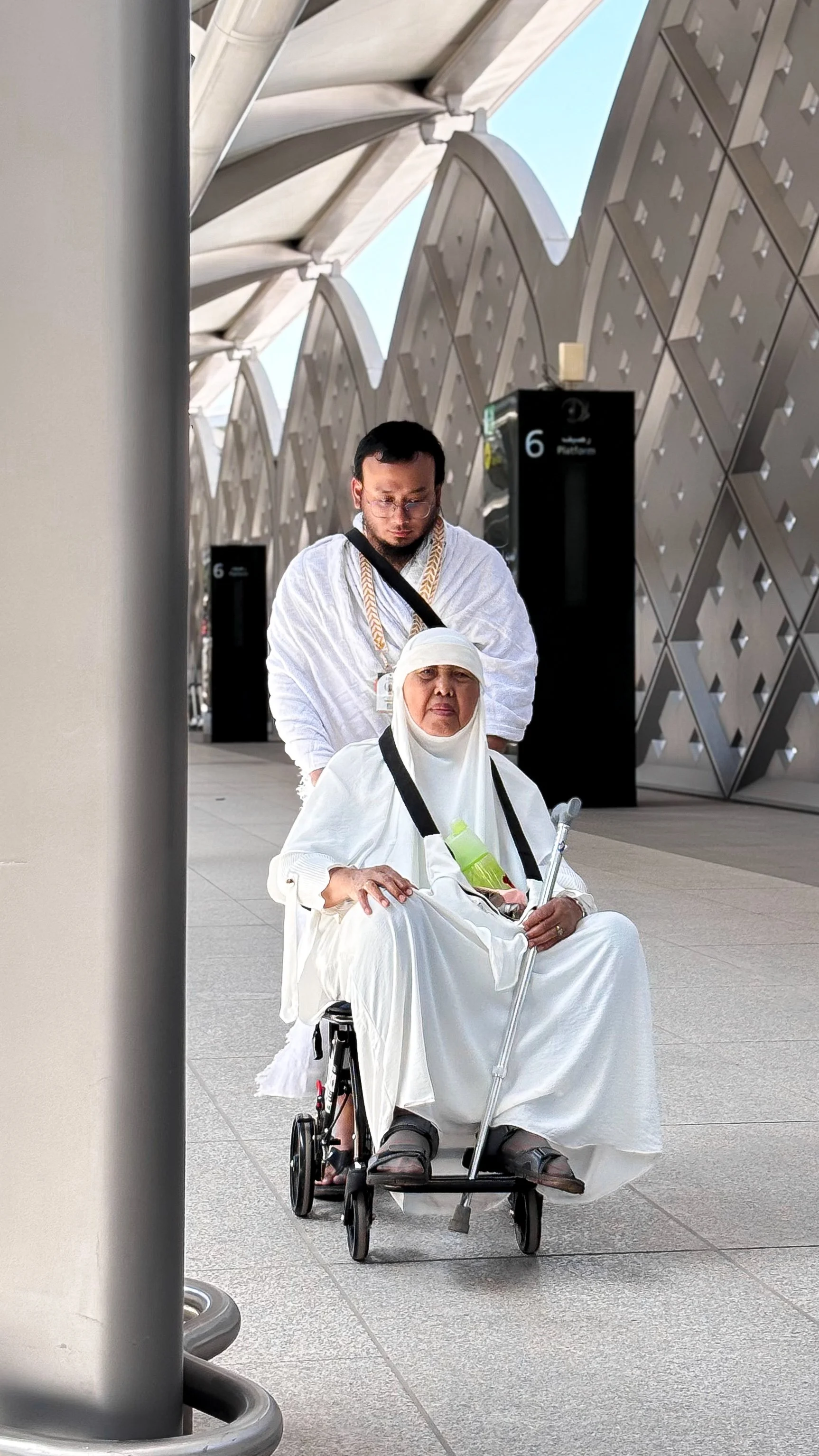 A woman dressed in white sitting in a wheelchair being pushed by a man also in white, both appear to be of Middle Eastern or South Asian descent, in an outdoor urban area.