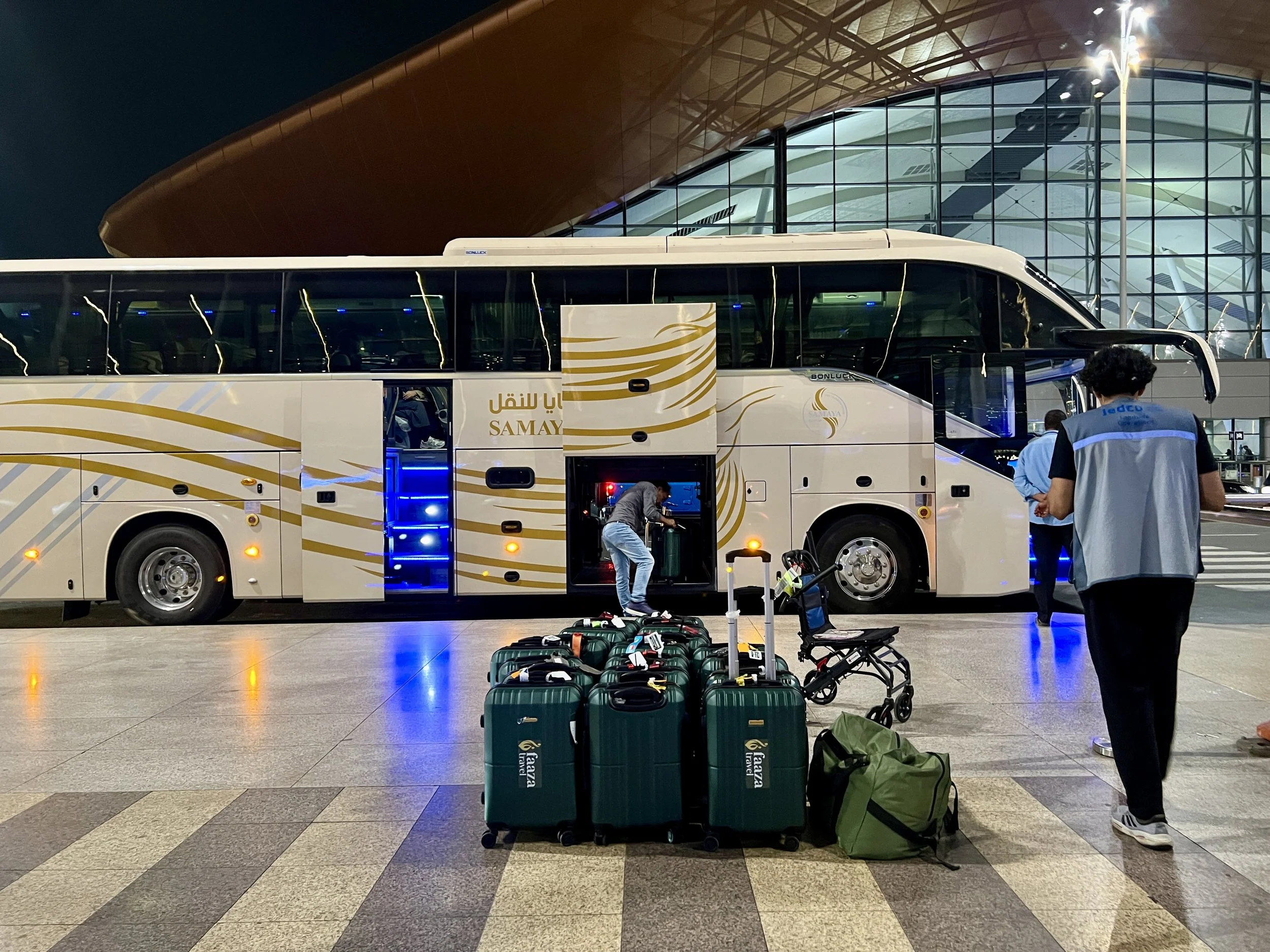 A white and yellow tour bus parked at an airport with baggage and a traveler with luggage. The airport terminal has large glass windows and modern architecture, illuminated by a tall street lamp, with a person working on luggage and others waiting nearby.