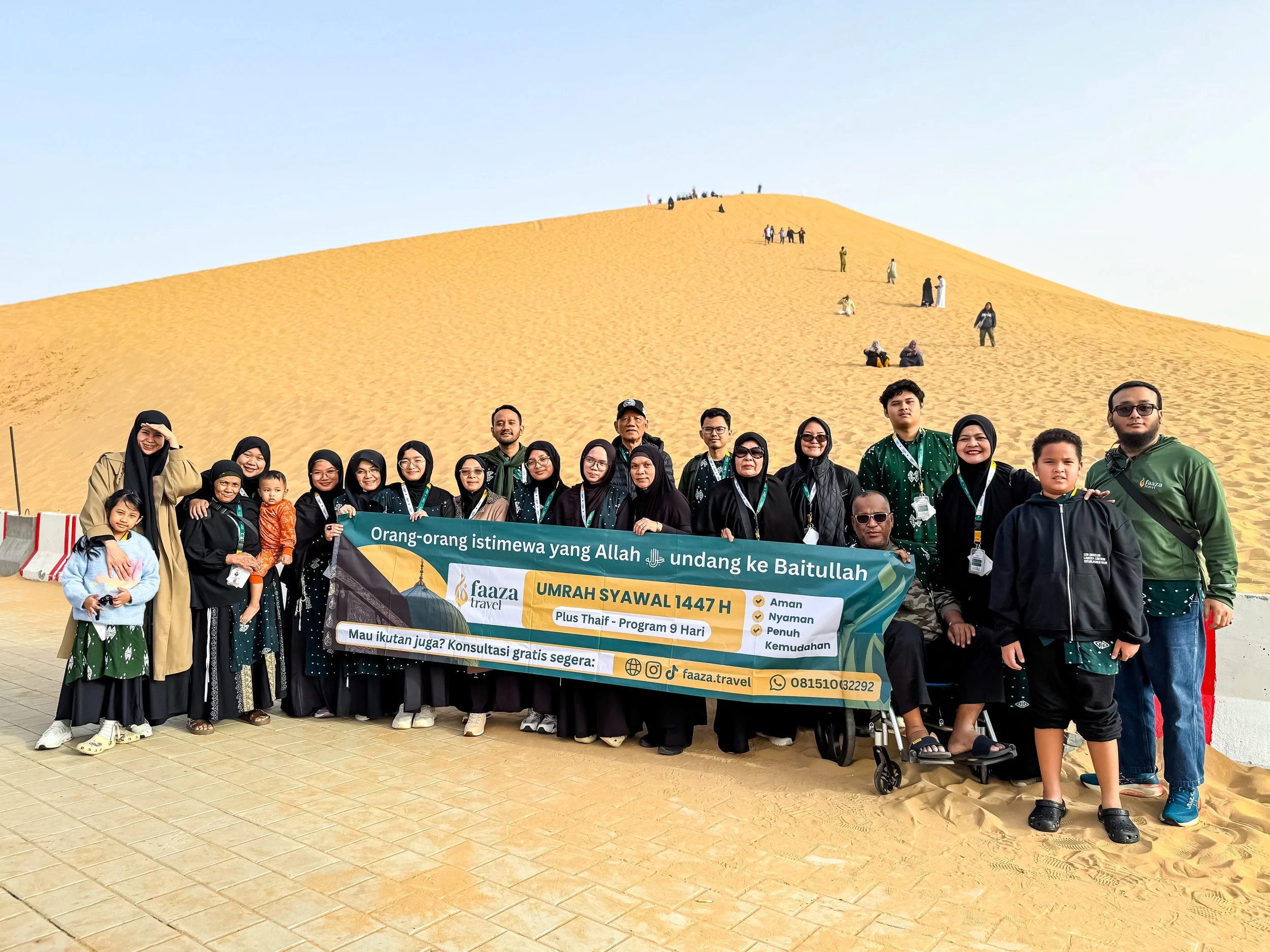 A group of people standing in front of a large sand dune, holding a banner advertising a travel program for Umrah during Eid al-Adha. The group includes men, women, children, and a person in a wheelchair. The background shows the sand dune with some 