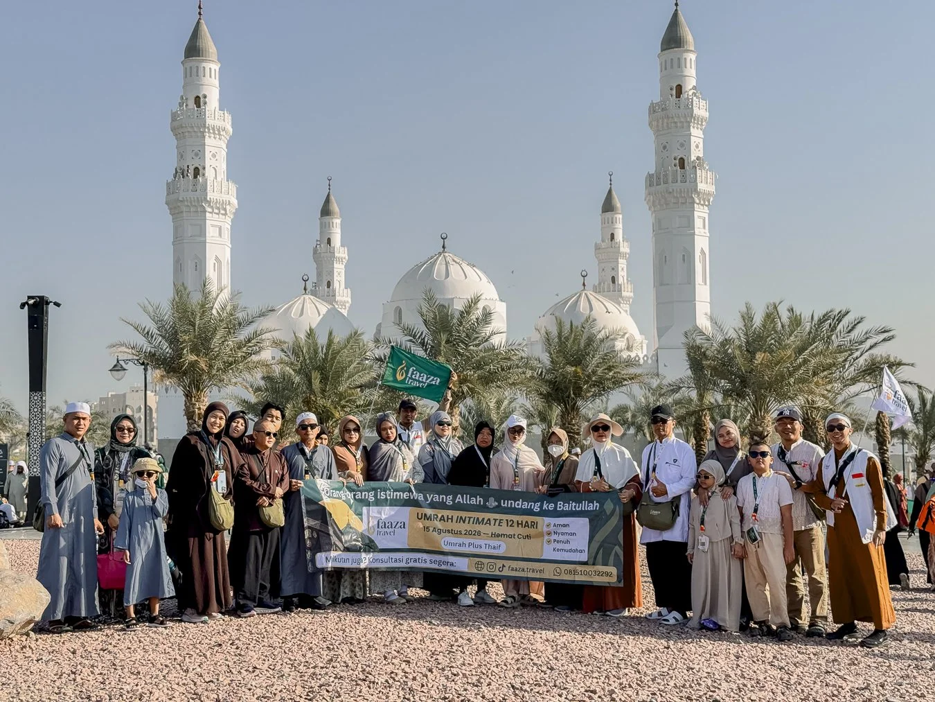 Group of people standing in front of a mosque with six minarets and white domes, holding a banner and flags, with palm trees and a clear sky in the background.