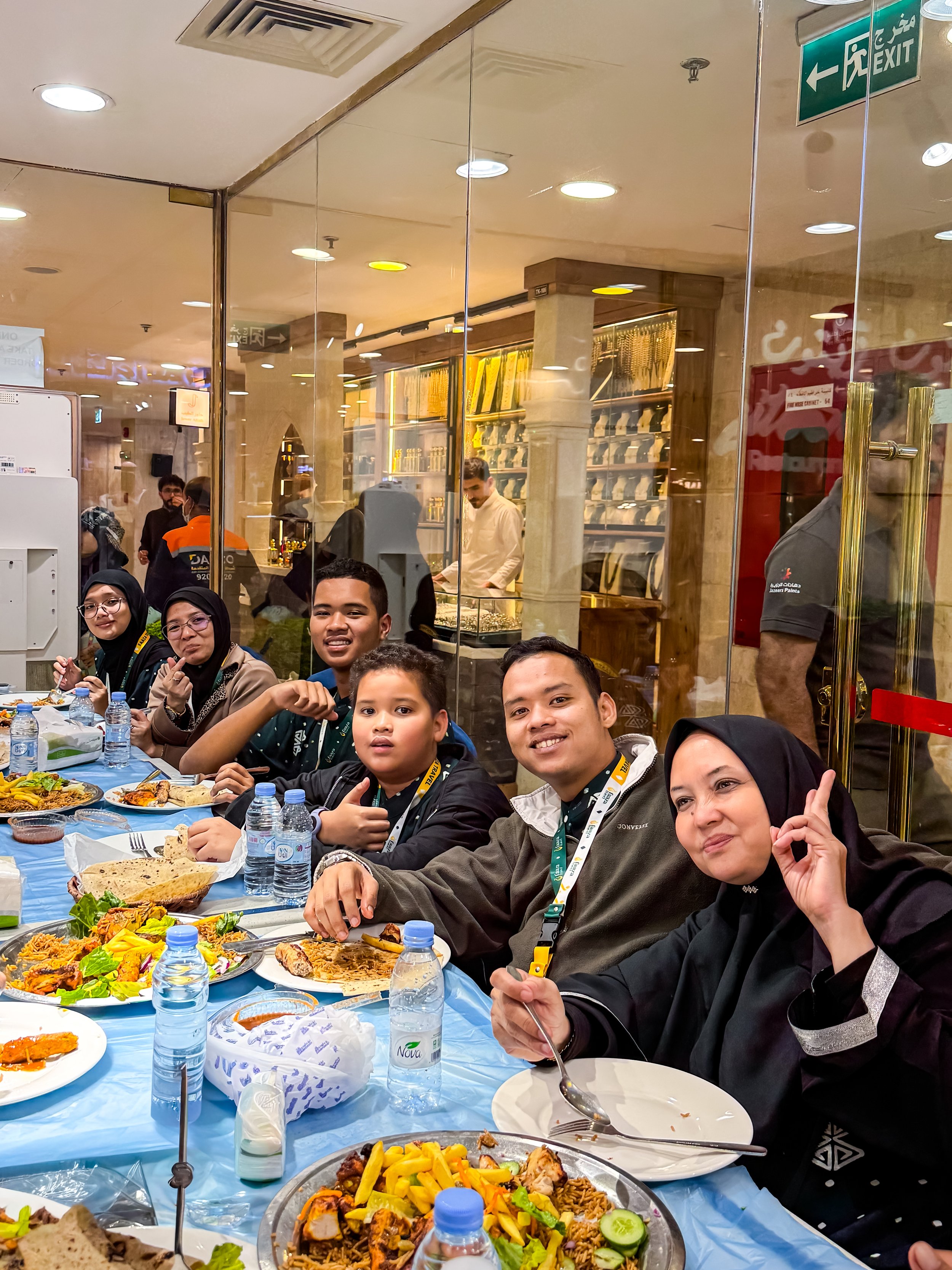 Group of people enjoying a meal at a restaurant, with traditional food dishes like rice, vegetables, and grilled meats on the table. The background reveals a display of shoes behind glass, and people working in the restaurant. The setting is indoors 
