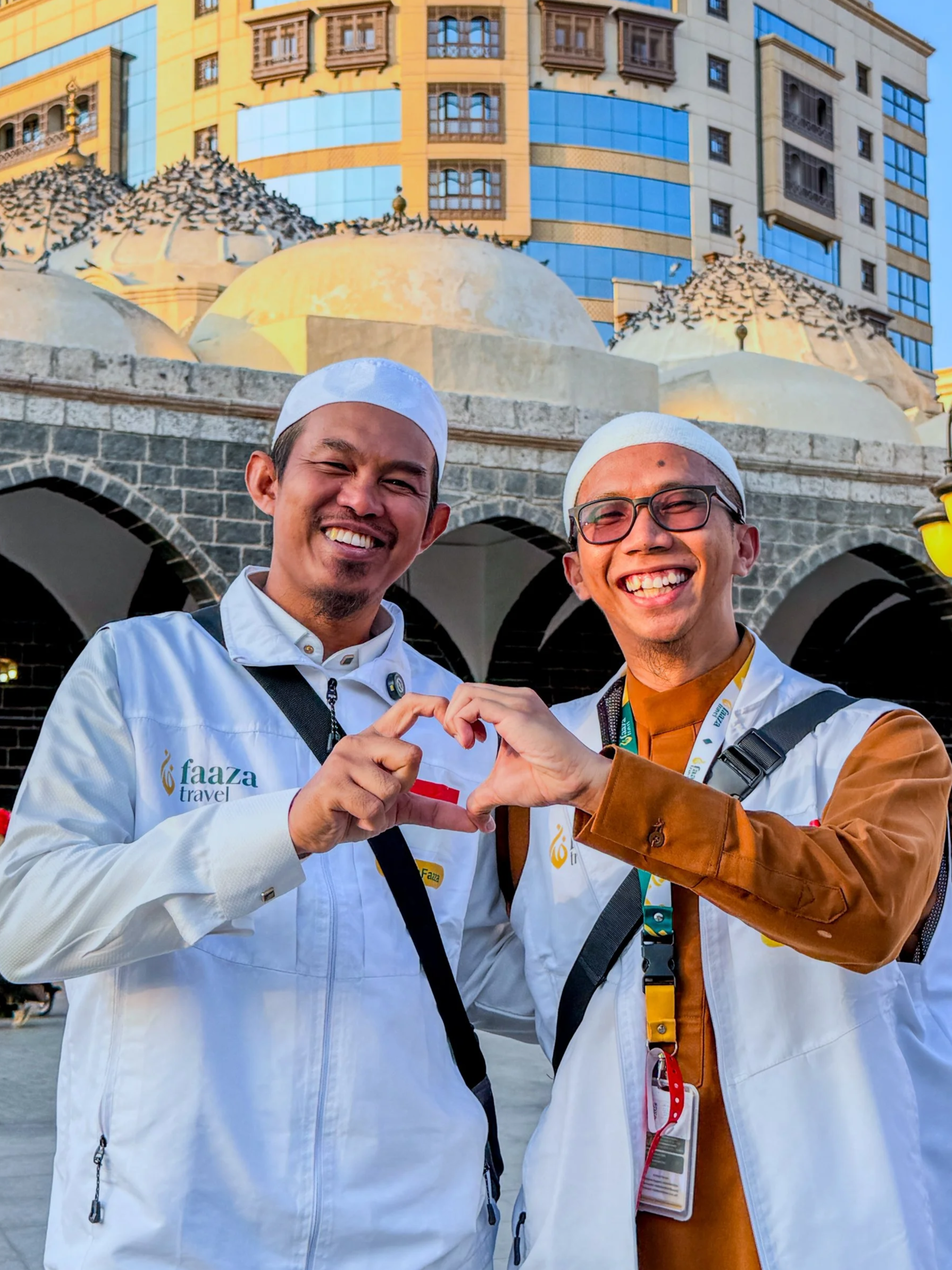 Two men smiling and forming a heart shape with their hands, standing outside with a large building in the background.