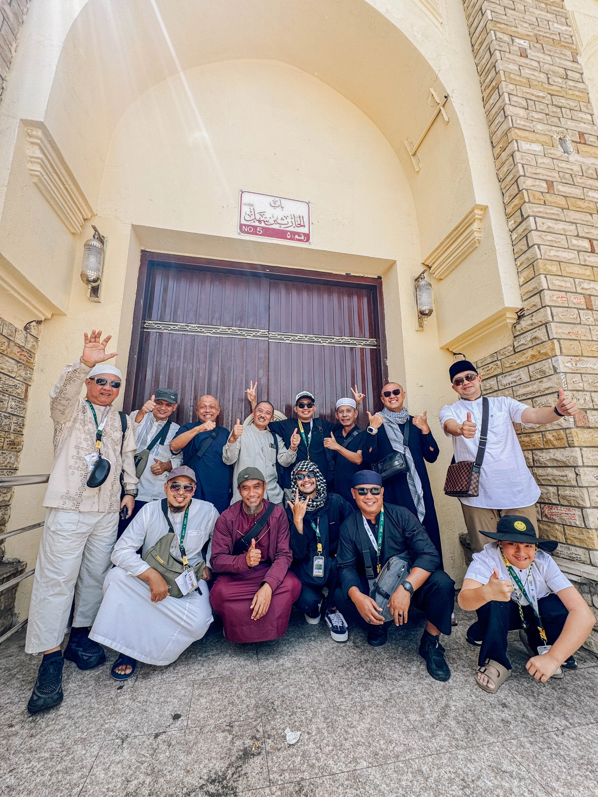 Group of smiling tourists posing in front of a large wooden gate and brick archway at a historic site.
