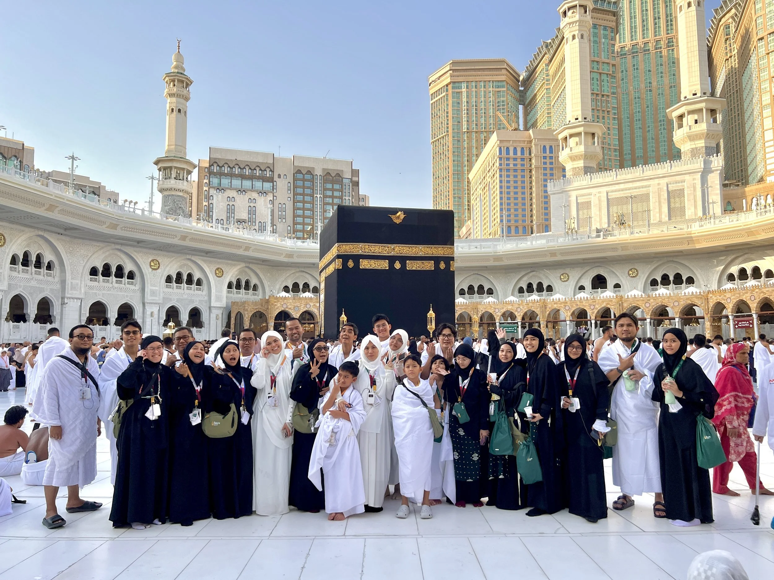 Group of people dressed in white and black clothing at the Masjid al-Haram in Mecca, Saudi Arabia, with the Kaaba at the center and modern buildings in the background.
