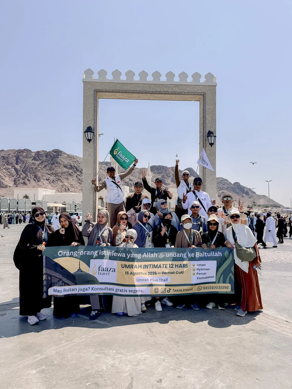 Group of people posing with a banner at the Masjid al-Haram in Mecca, Saudi Arabia, with a large empty frame structure and mountains in the background during daytime.
