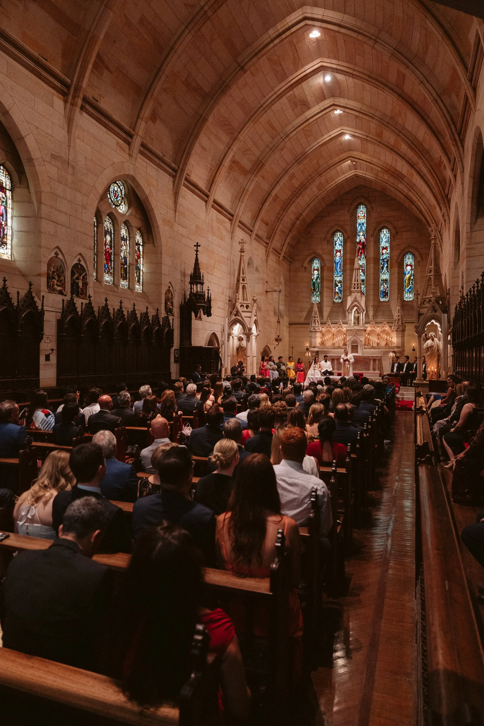 A large gathering of people attending a wedding ceremony inside a church with stained glass windows and high vaulted ceilings.