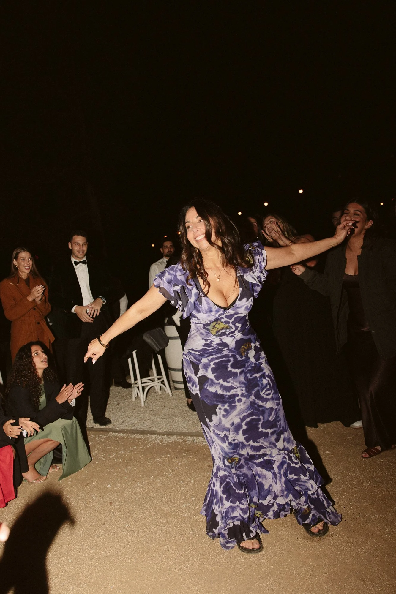 Woman dancing in a purple floral dress at an outdoor evening event, with people watching and enjoying her dance.