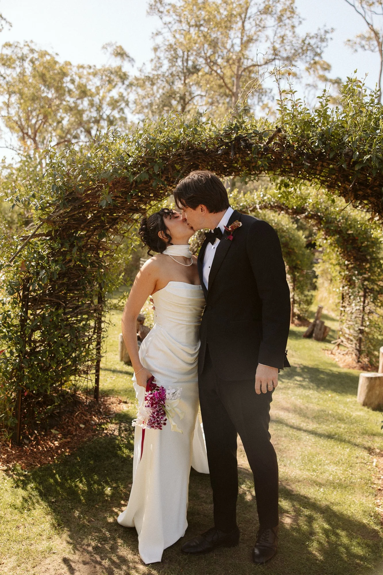 A bride and groom kissing beneath a green arch in an outdoor garden setting during their wedding ceremony.