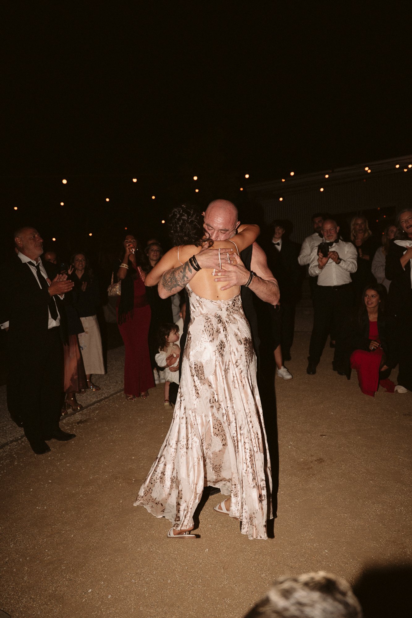 A couple sharing a dance at their wedding, surrounded by guests in an outdoor setting at night with string lights overhead.