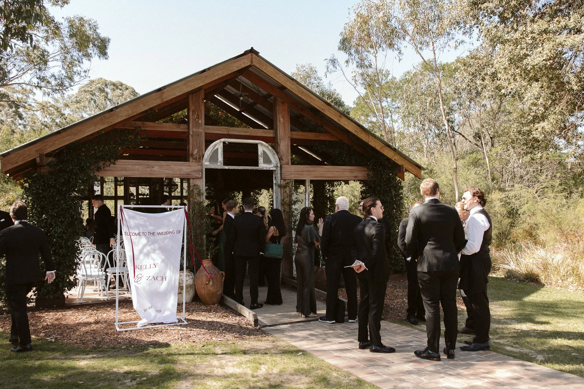 People dressed in formal attire gathering outdoors at a wedding reception in front of a rustic wooden building with trees and greenery in the background.