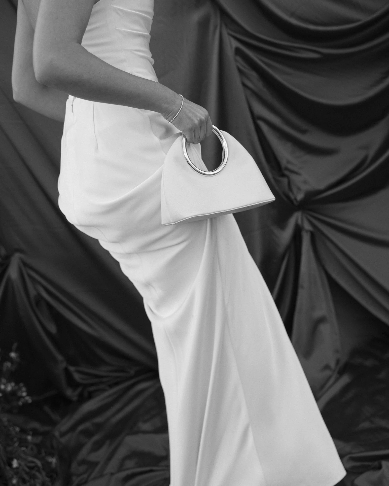 A woman in a white dress holding a white handbag with circular handles, standing against a dark fabric backdrop.