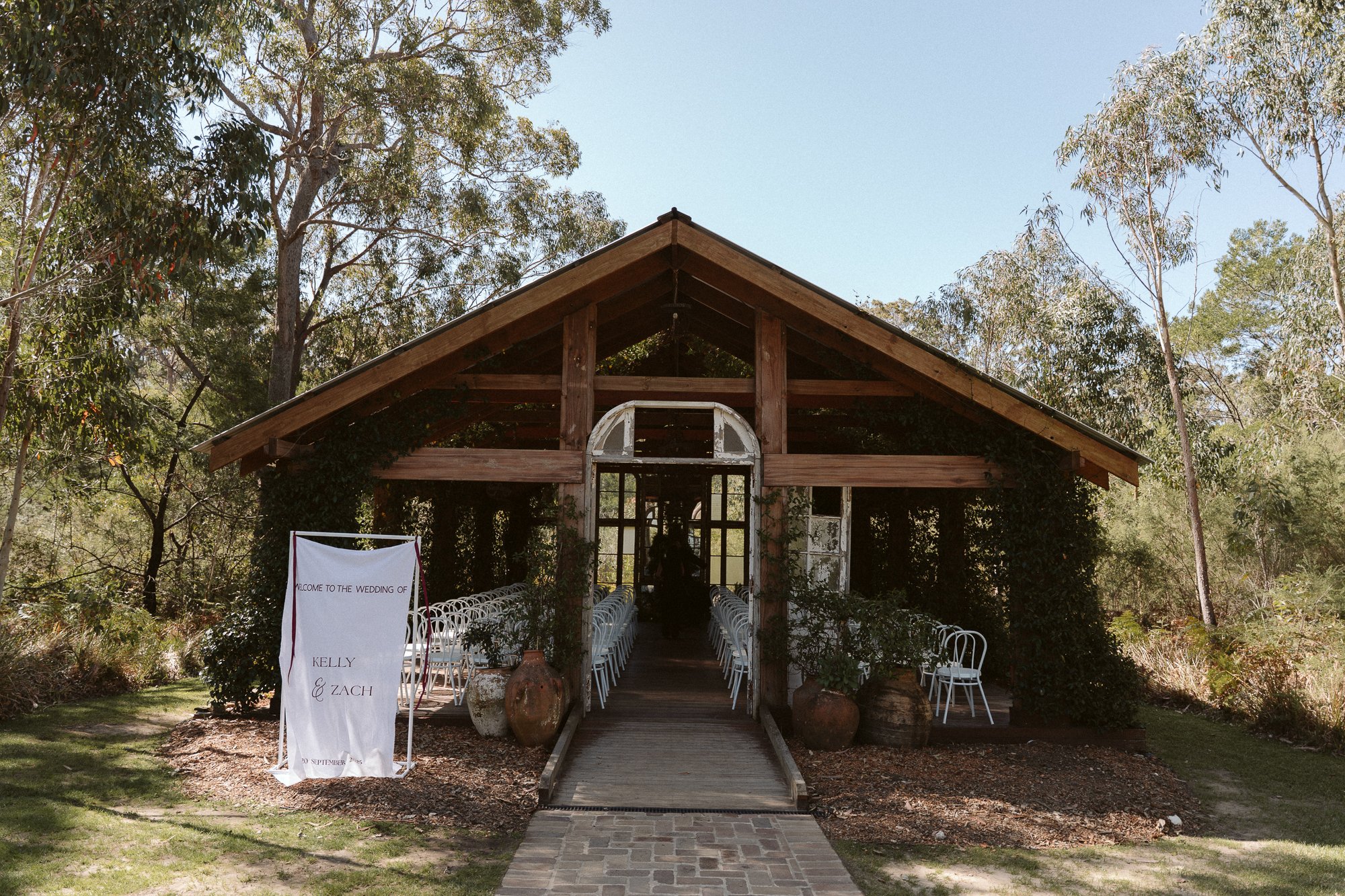 Wooden outdoor wedding venue with white chairs, a sign welcoming to Kelly and Zach's wedding, surrounded by trees and greenery.