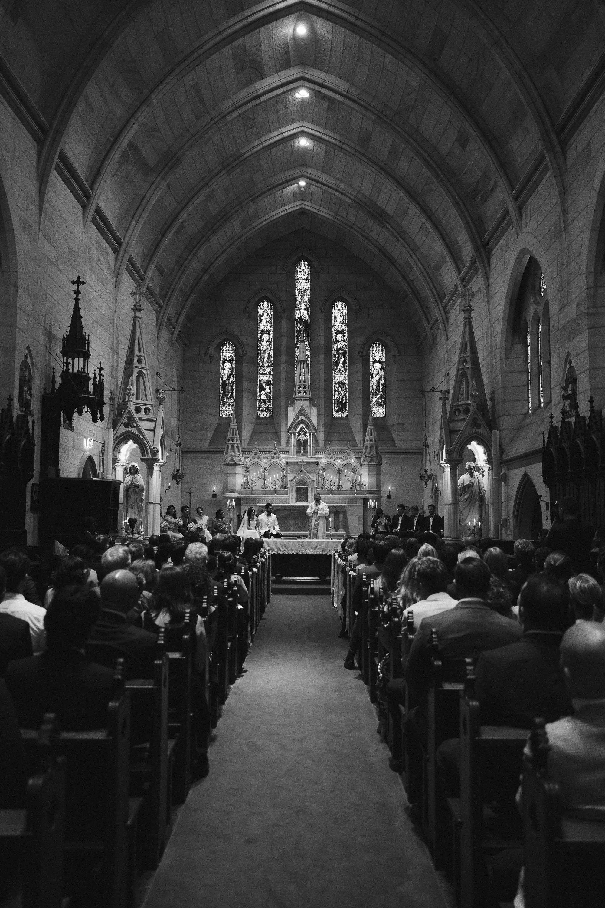 Black and white photo of a church interior during a wedding ceremony with guests seated, altar at the front, stained glass windows, and decorative statues.
