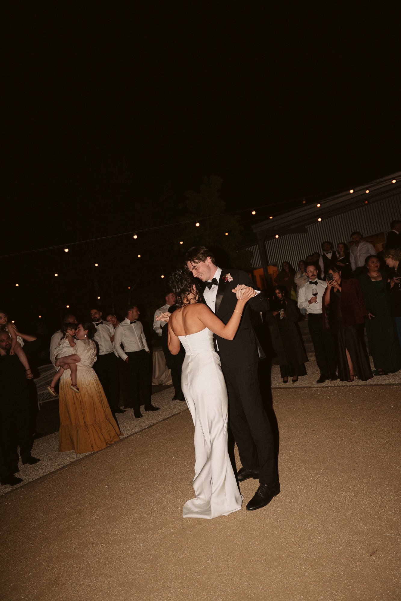 A bride and groom dance at their wedding reception at night, surrounded by guests. The bride wears a white strapless dress, and the groom is in a black tuxedo. String lights hang overhead in an outdoor setting.
