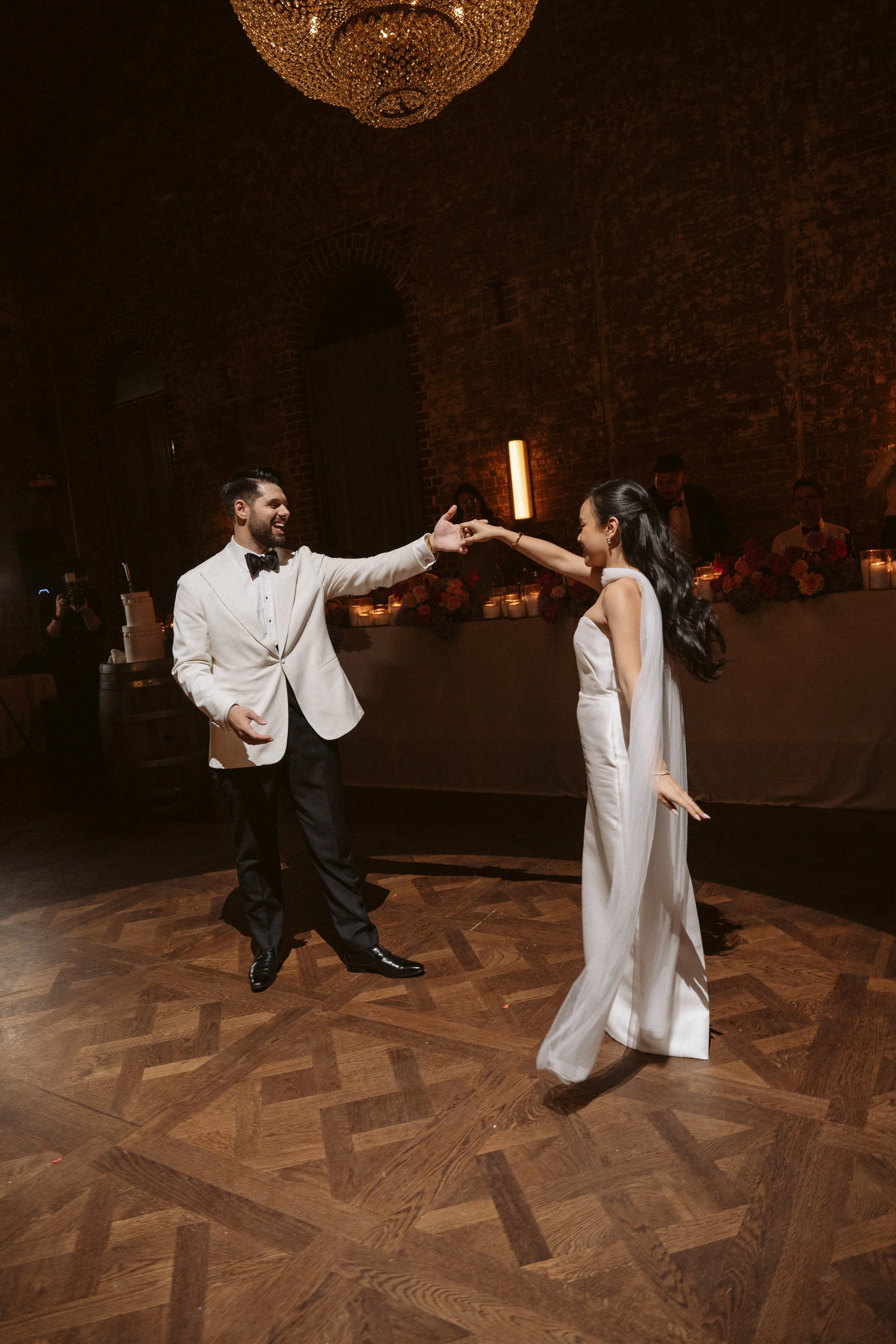A couple dancing at a wedding reception, with the groom in a white tuxedo jacket and the bride in a white gown, in a dimly lit venue with a chandelier above.