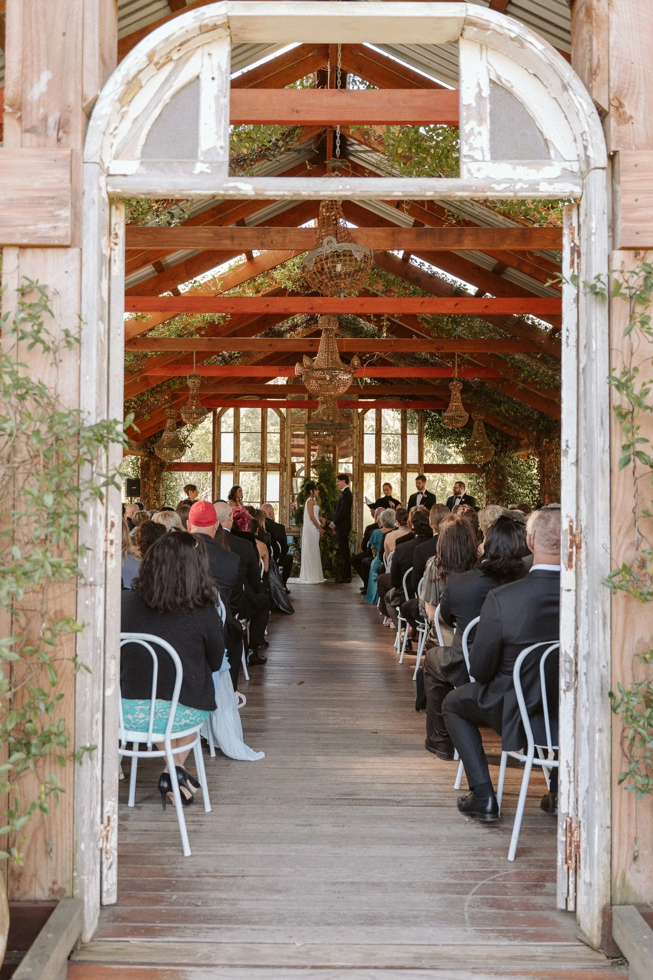 Wedding ceremony inside a rustic, greenhouse-style venue with wooden beams, chandeliers, and large windows, viewed through a weathered wooden doorway.