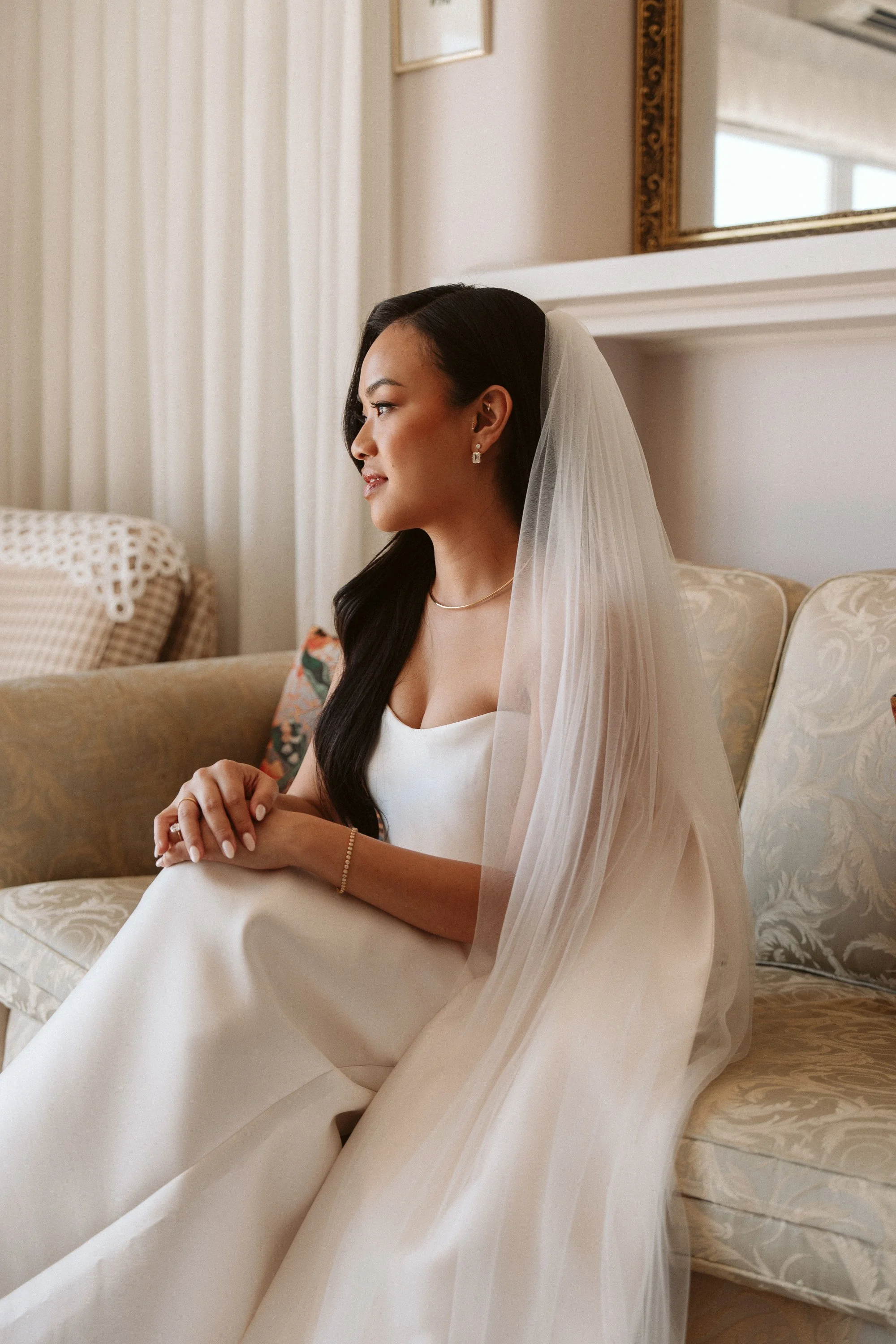 A bride sitting on a vintage sofa, dressed in a white wedding gown with a veiled hairstyle, jewelry, and a calm expression, inside a softly lit room.