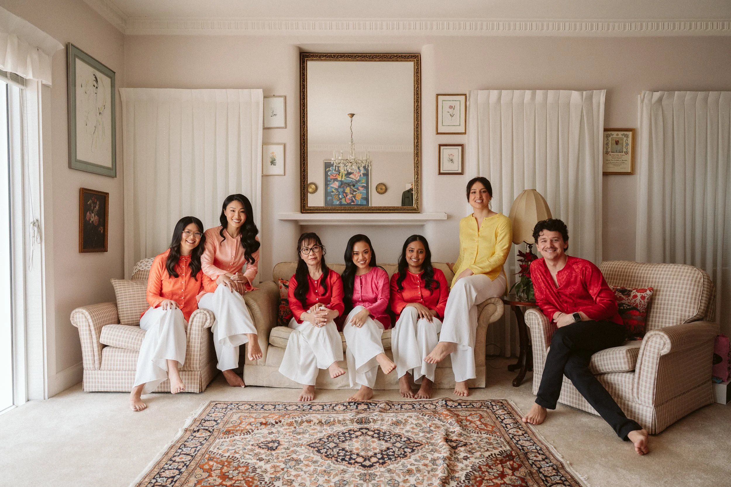 A group of eight people, consisting of women and men, sitting and standing in a living room. They are dressed in colorful traditional clothing and are smiling. The room has a mirror, framed pictures, a sofa, an armchair, and a patterned rug.