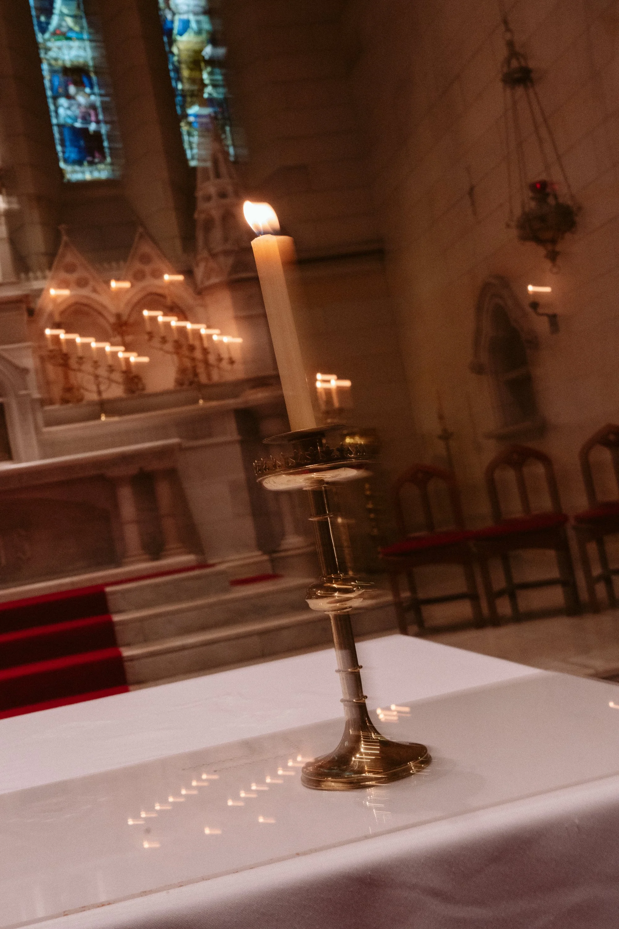 A lit candle on a brass candlestick holder placed on a white altar cloth inside a church, with stained glass windows and wooden chairs in the background.