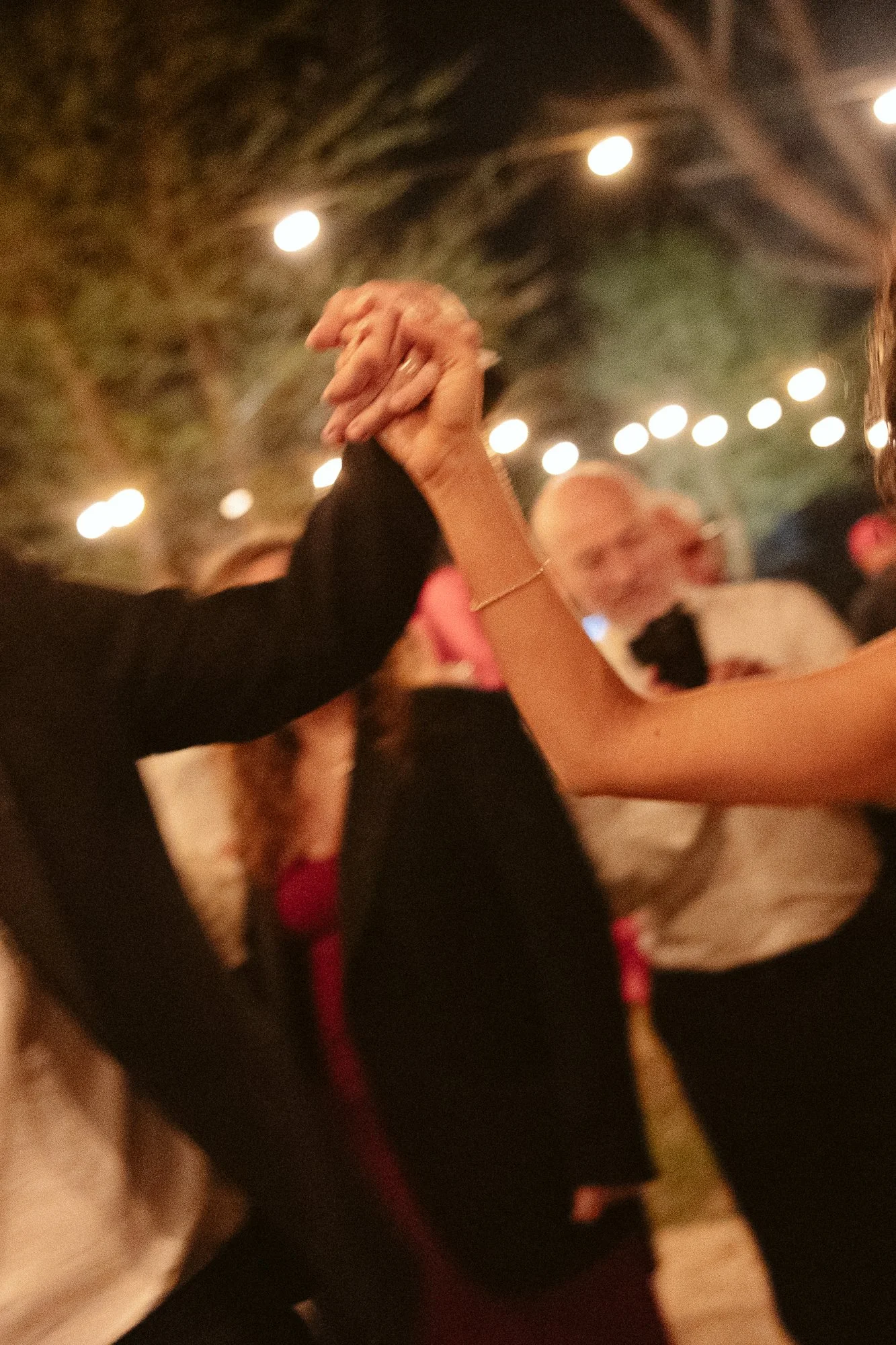 People holding hands while dancing at an outdoor evening event with string lights overhead.