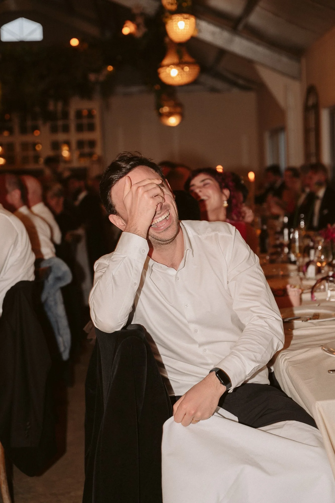 A man in a white shirt laughing or smiling while covering his face, seated at a banquet table with other people in the background at a formal event or celebration.