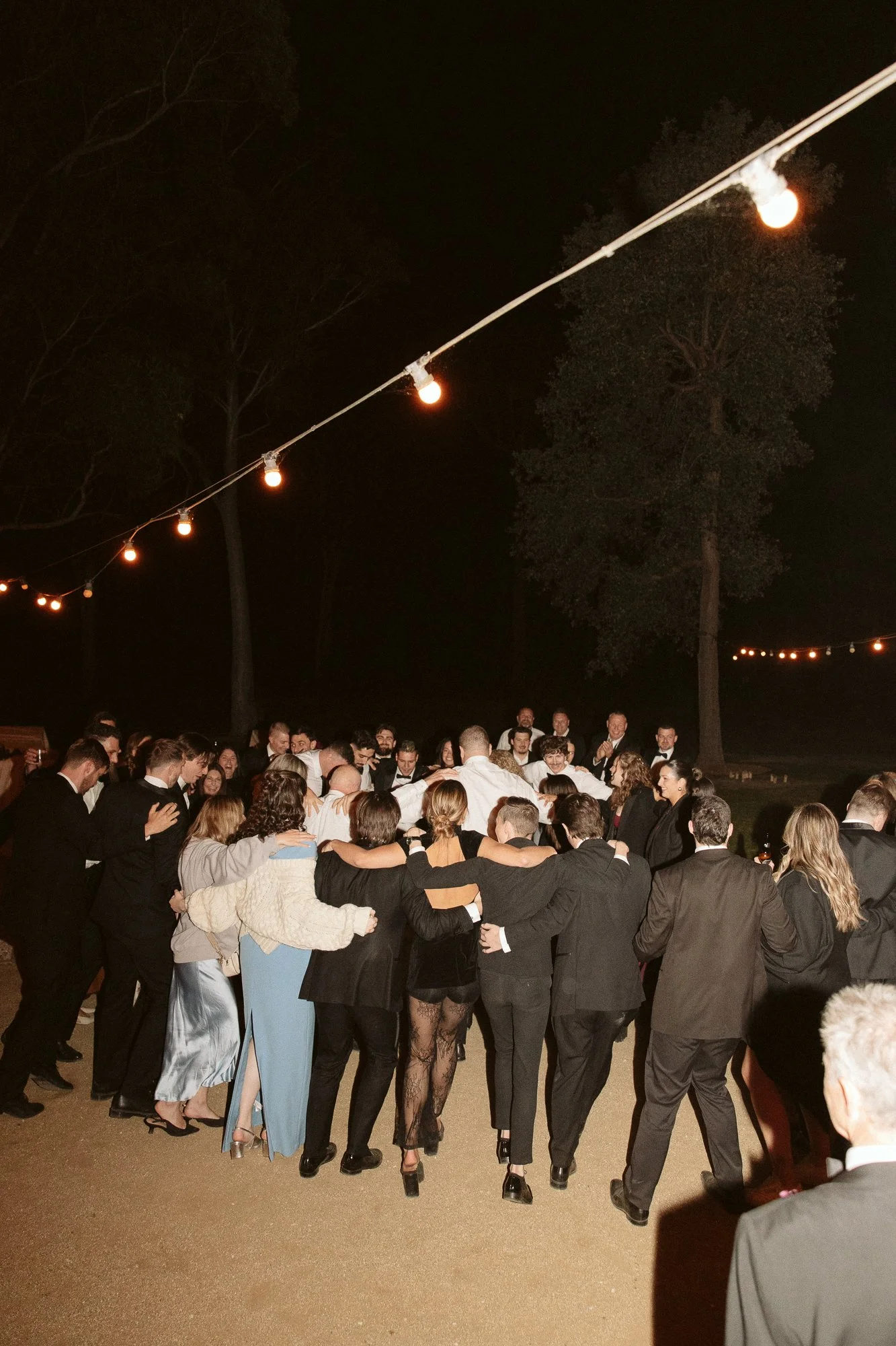 A large group of people dressed in formal attire, gathered outdoors at night under string lights, participating in a group hug or dance circle at a celebration or wedding.