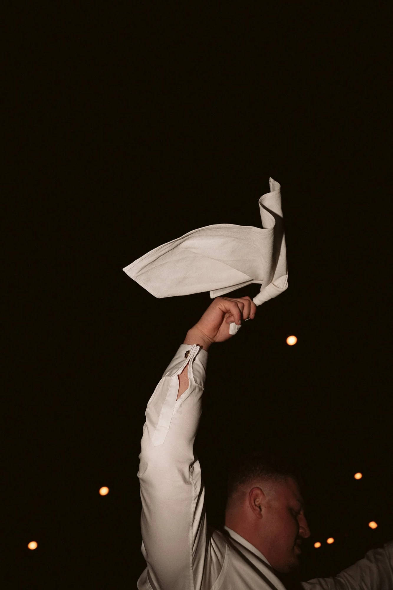 Person waving a white cloth at night, with a dark sky and small orange lights in the background.