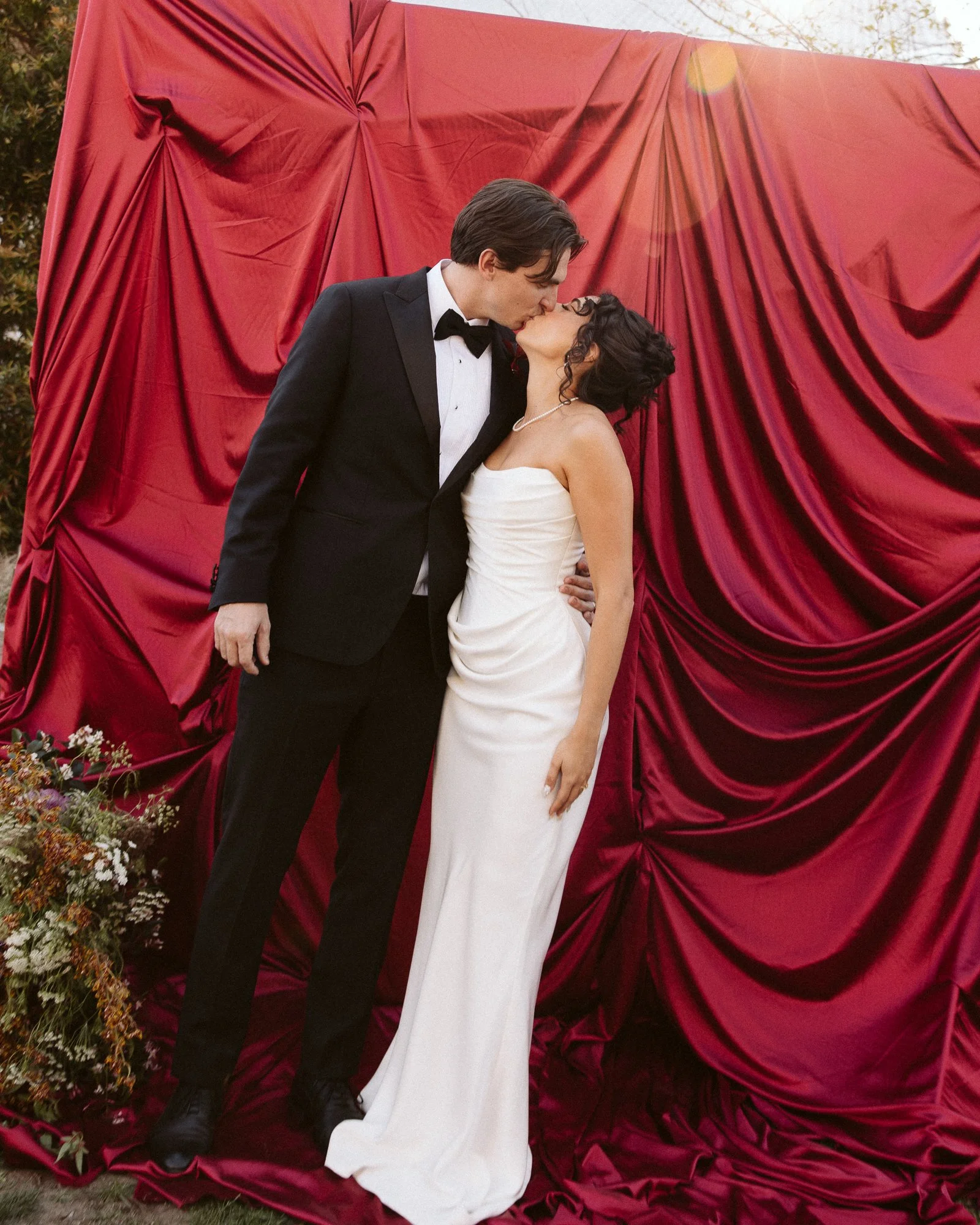 A couple dressed in wedding attire kissing in front of a red draped backdrop, with a flower arrangement on the side.