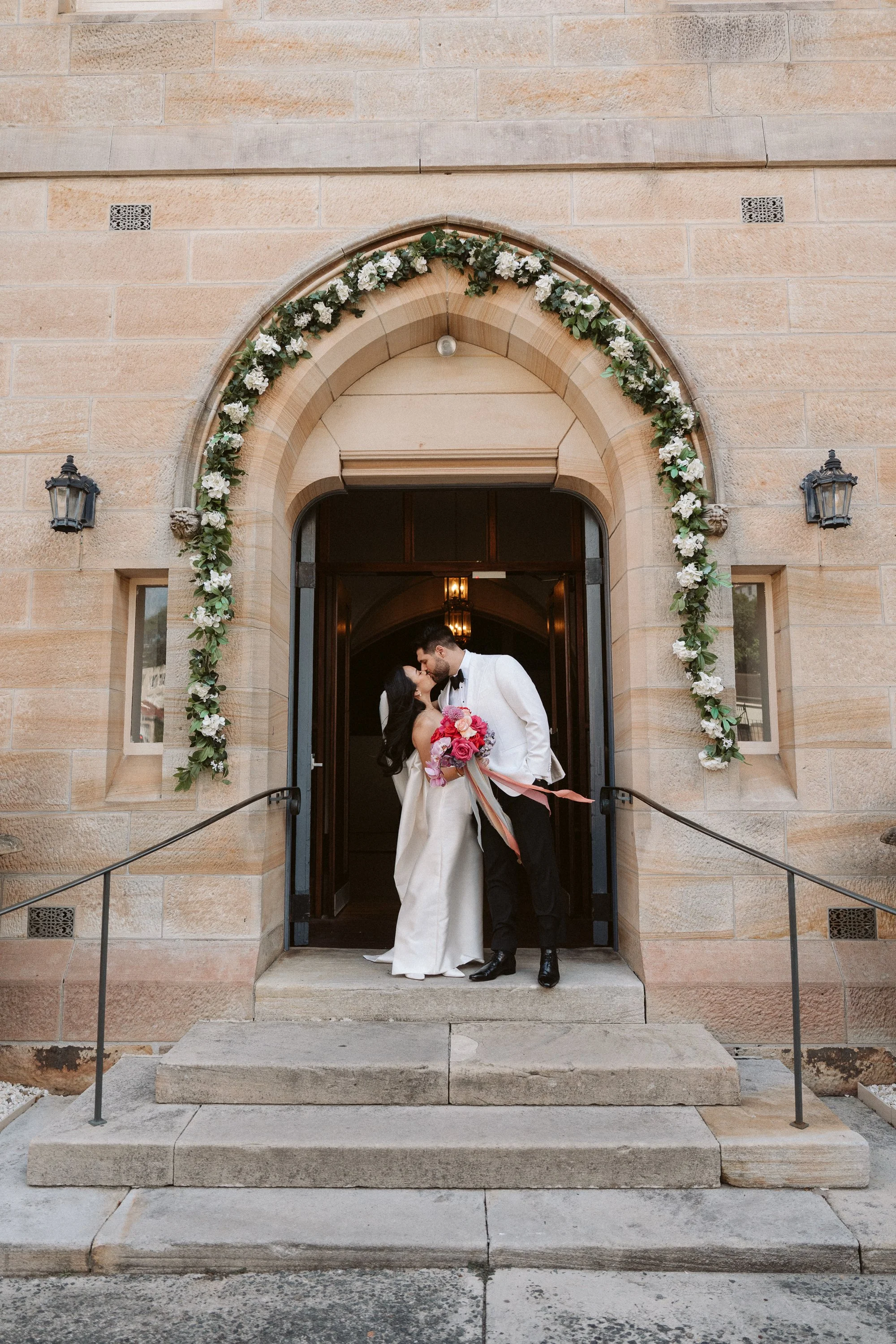 A newlywed couple sharing a kiss at the front entrance of a church, decorated with a floral arch with white flowers and greenery, during their wedding ceremony.