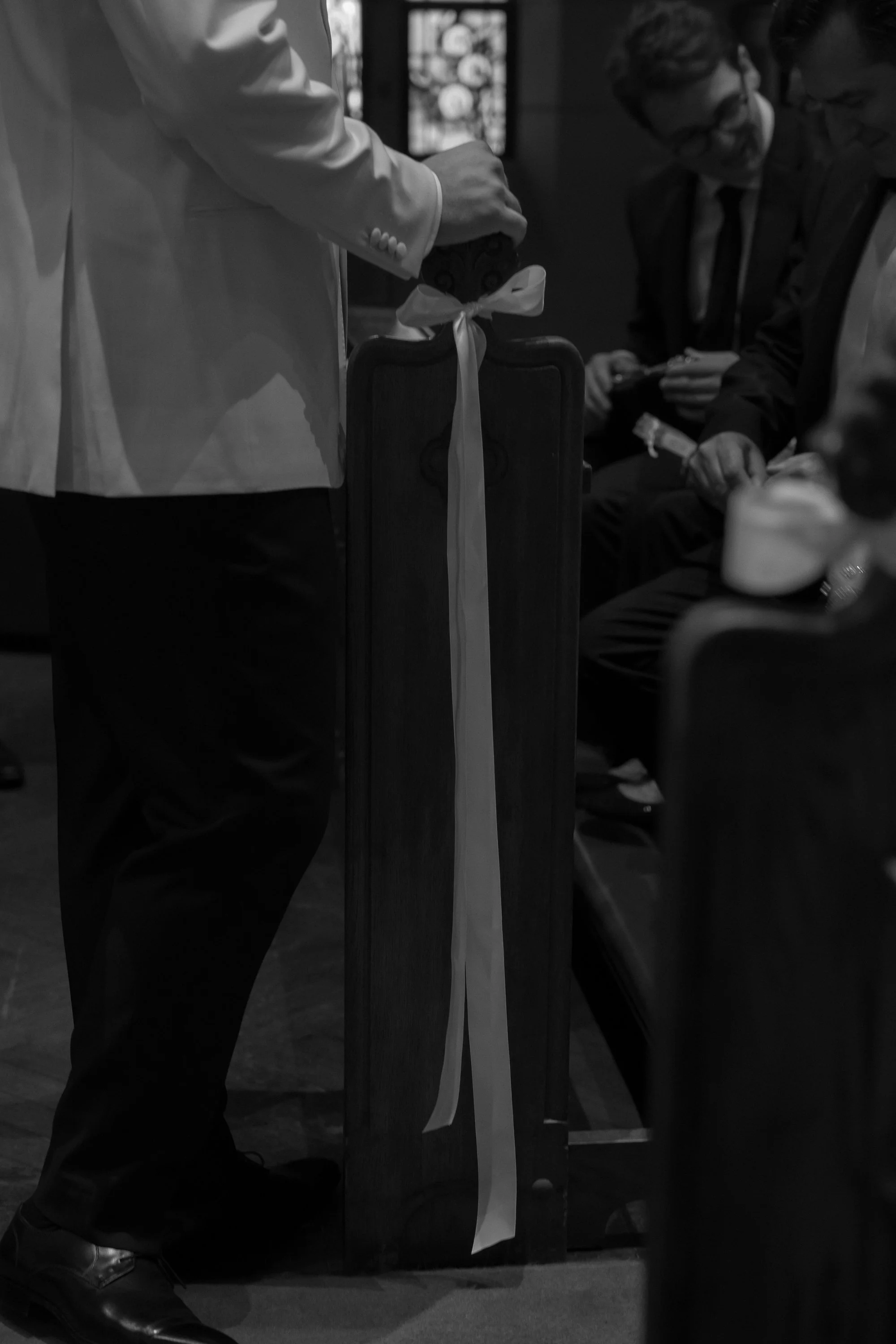 People sitting in chairs in a religious or ceremonial setting, with a wooden stand decorated with a ribbon and a person standing nearby.