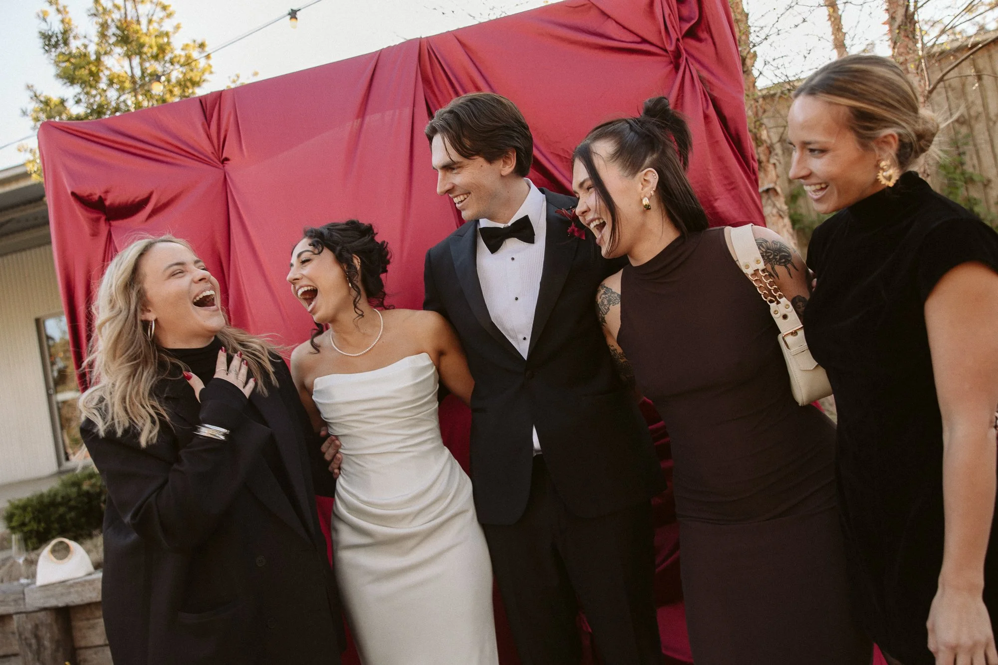 A group of five people, three women and two men, dressed in formal attire, standing outdoors in front of a red backdrop, laughing and enjoying themselves at a celebration or special event.