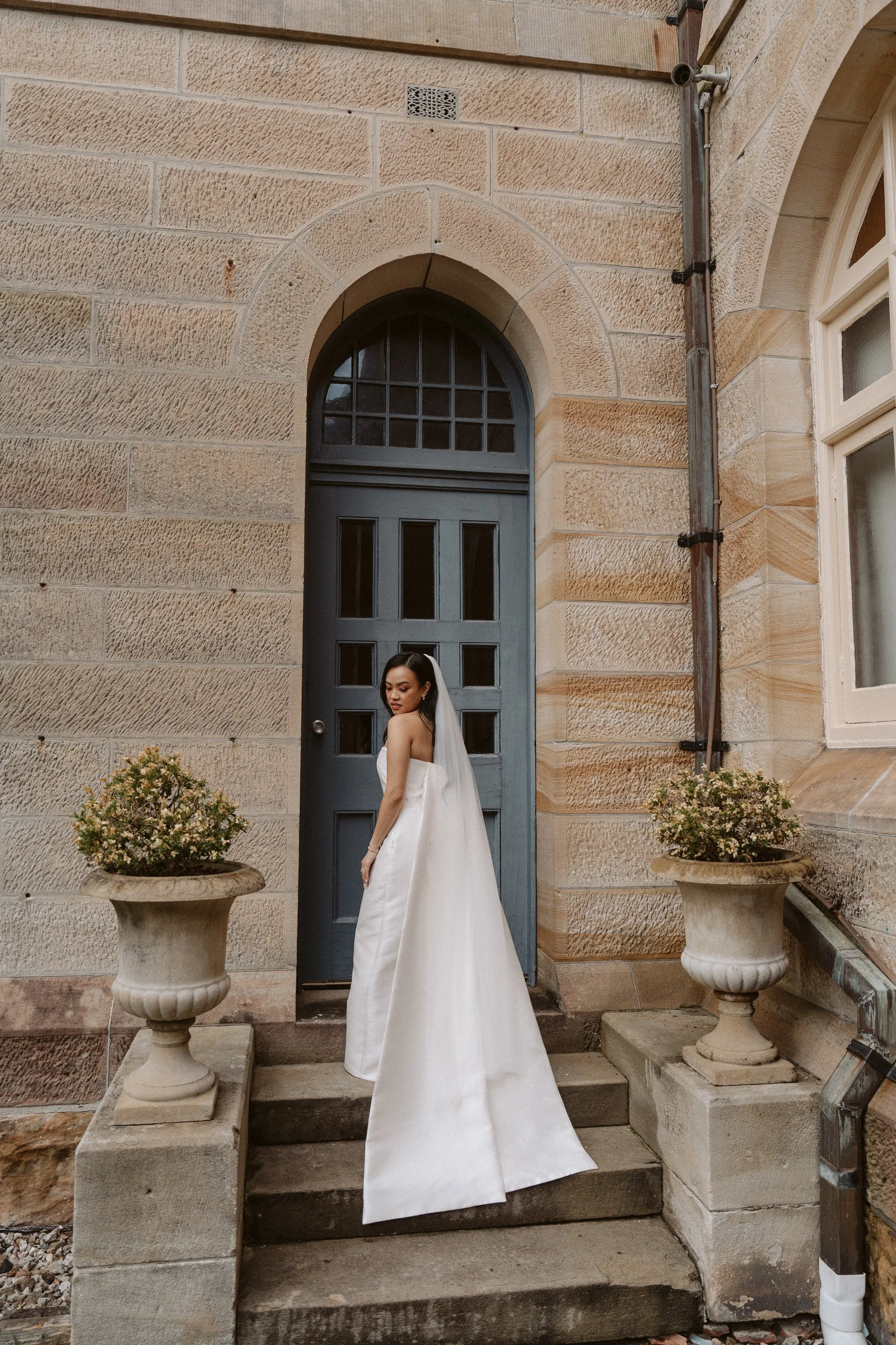 A woman in a white strapless wedding dress and veil standing on stone steps in front of a dark gray door, flanked by two large stone planters with flowers, outside a brick building.
