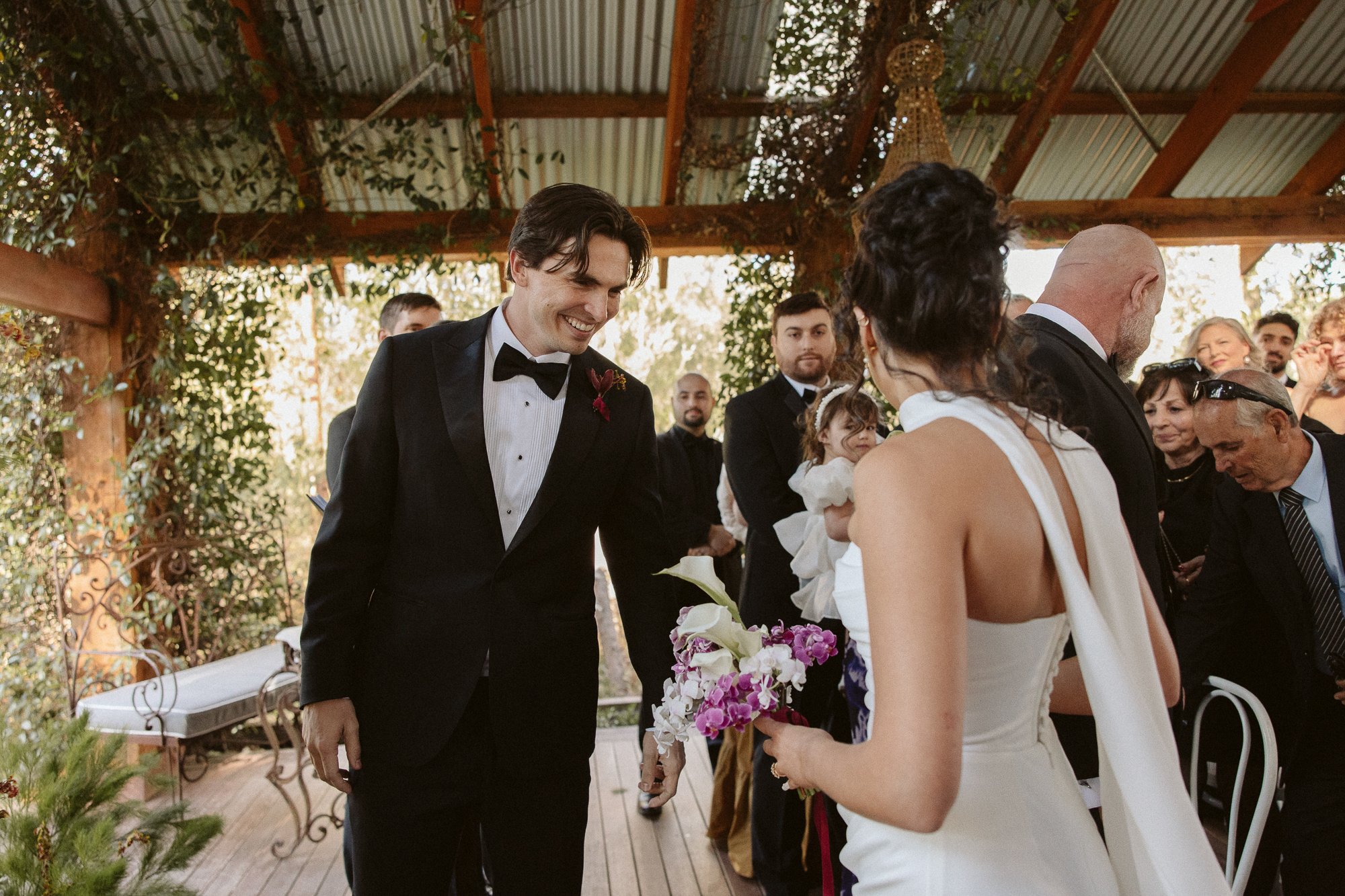 A wedding ceremony with a groom smiling at his bride, who is holding a bouquet of flowers. The bride is dressed in a white gown with a long veil. Other guests are present, some standing and watching, in a venue with wooden beams and greenery.