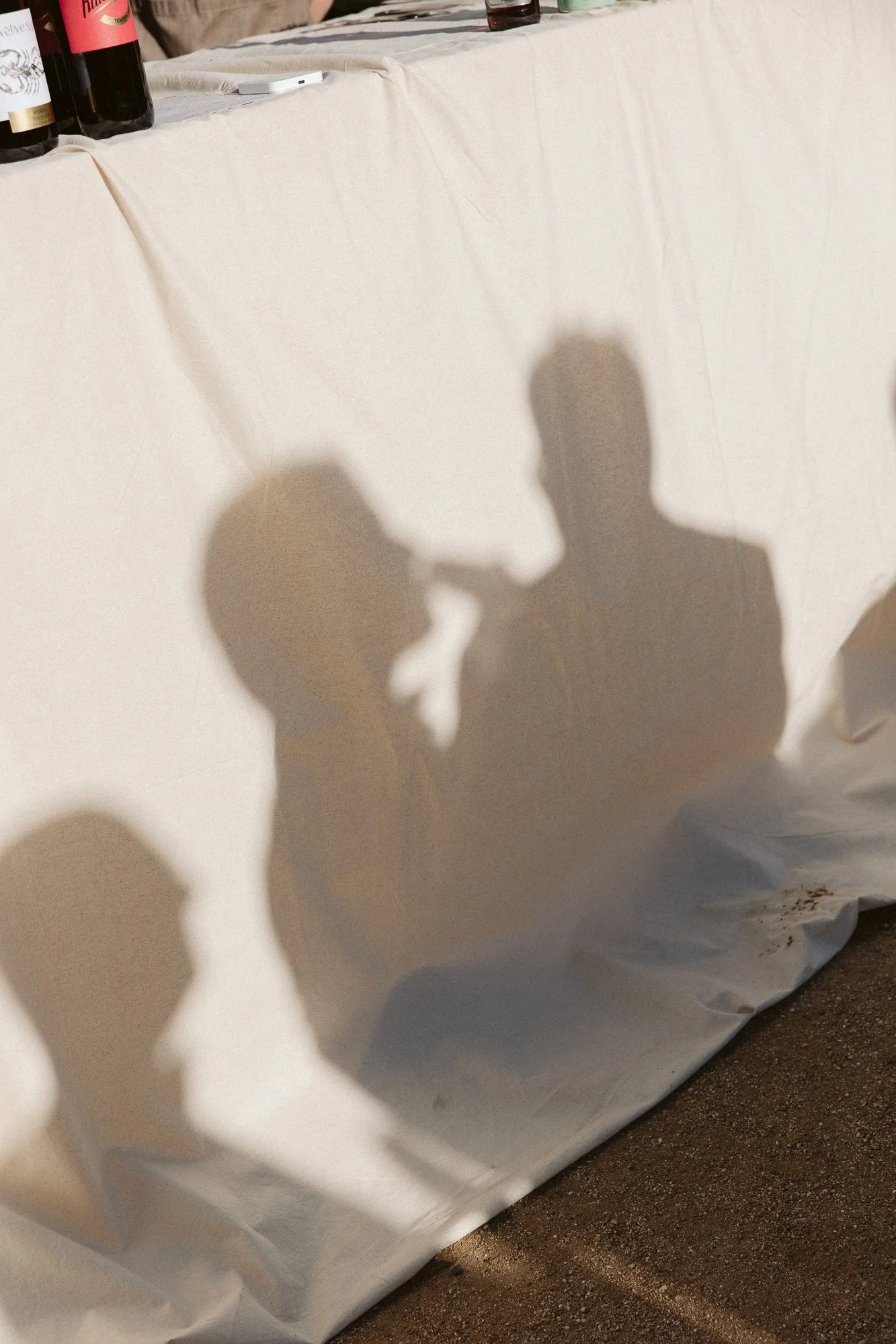 Shadows of two people on a white tablecloth with bottles of wine and other items on top.
