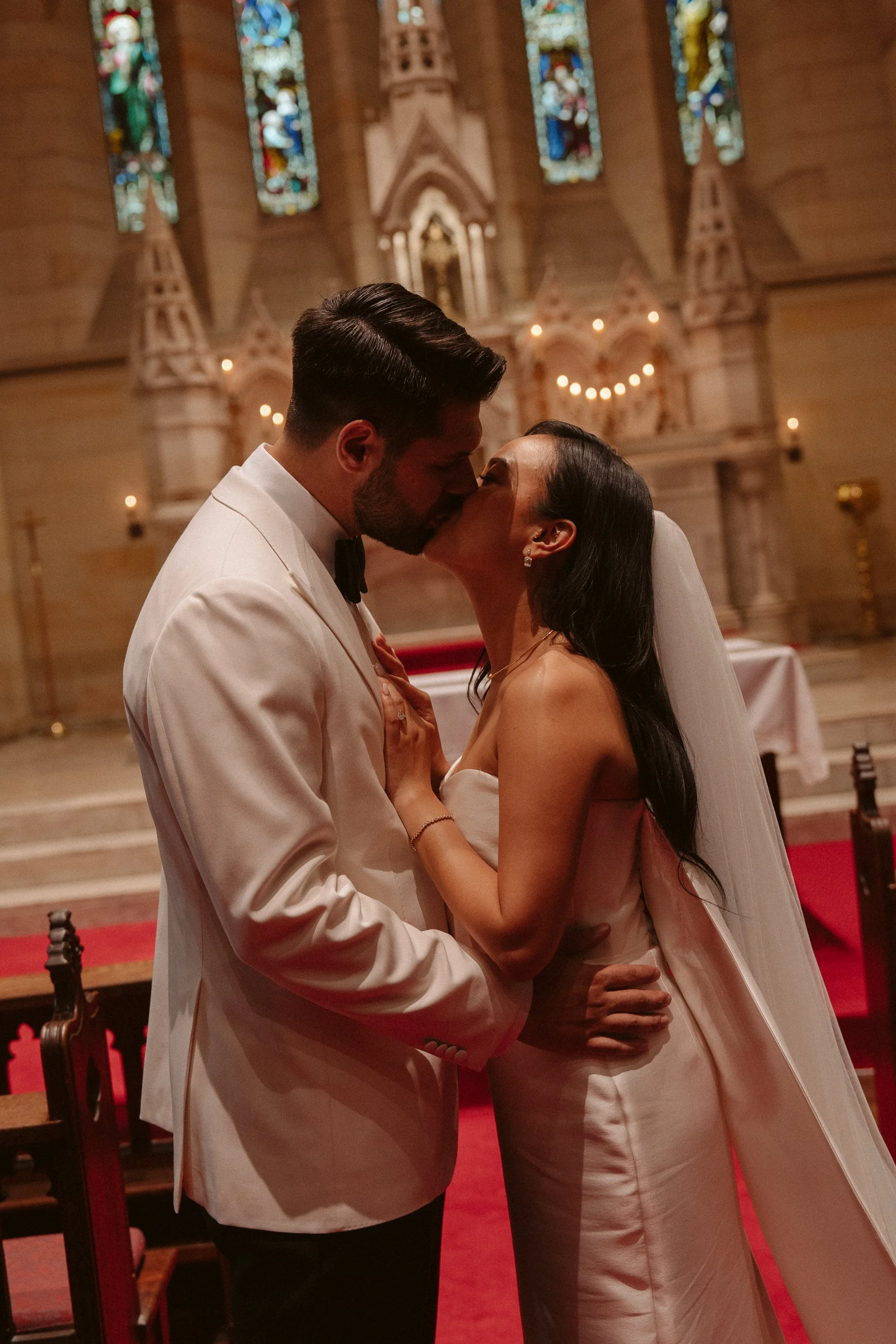 A newlywed couple sharing a kiss inside a church, with stained glass windows and religious decor in the background.