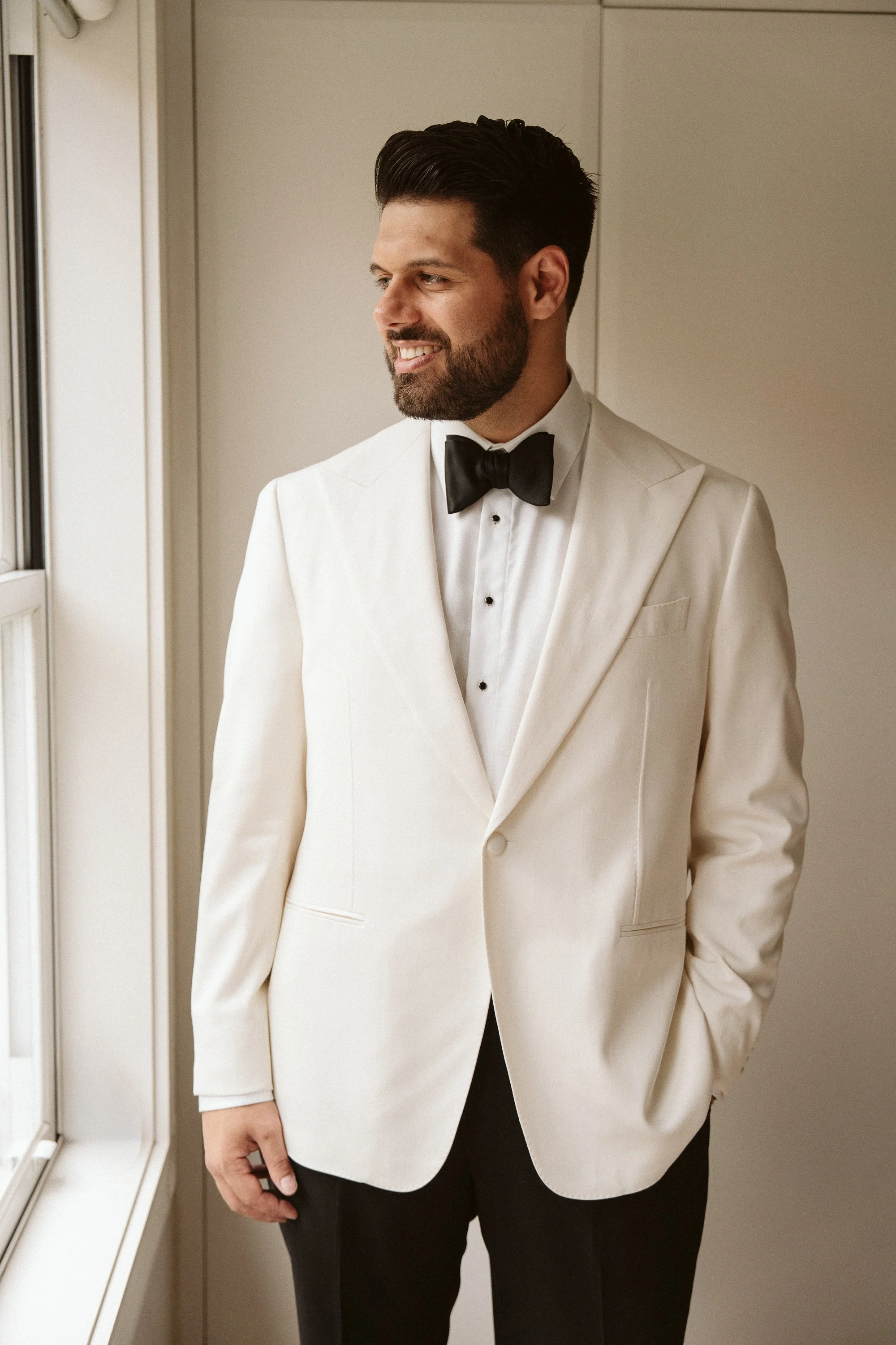 A man wearing a white tuxedo, black bow tie, and black pants standing near a window, looking outside with a slight smile.