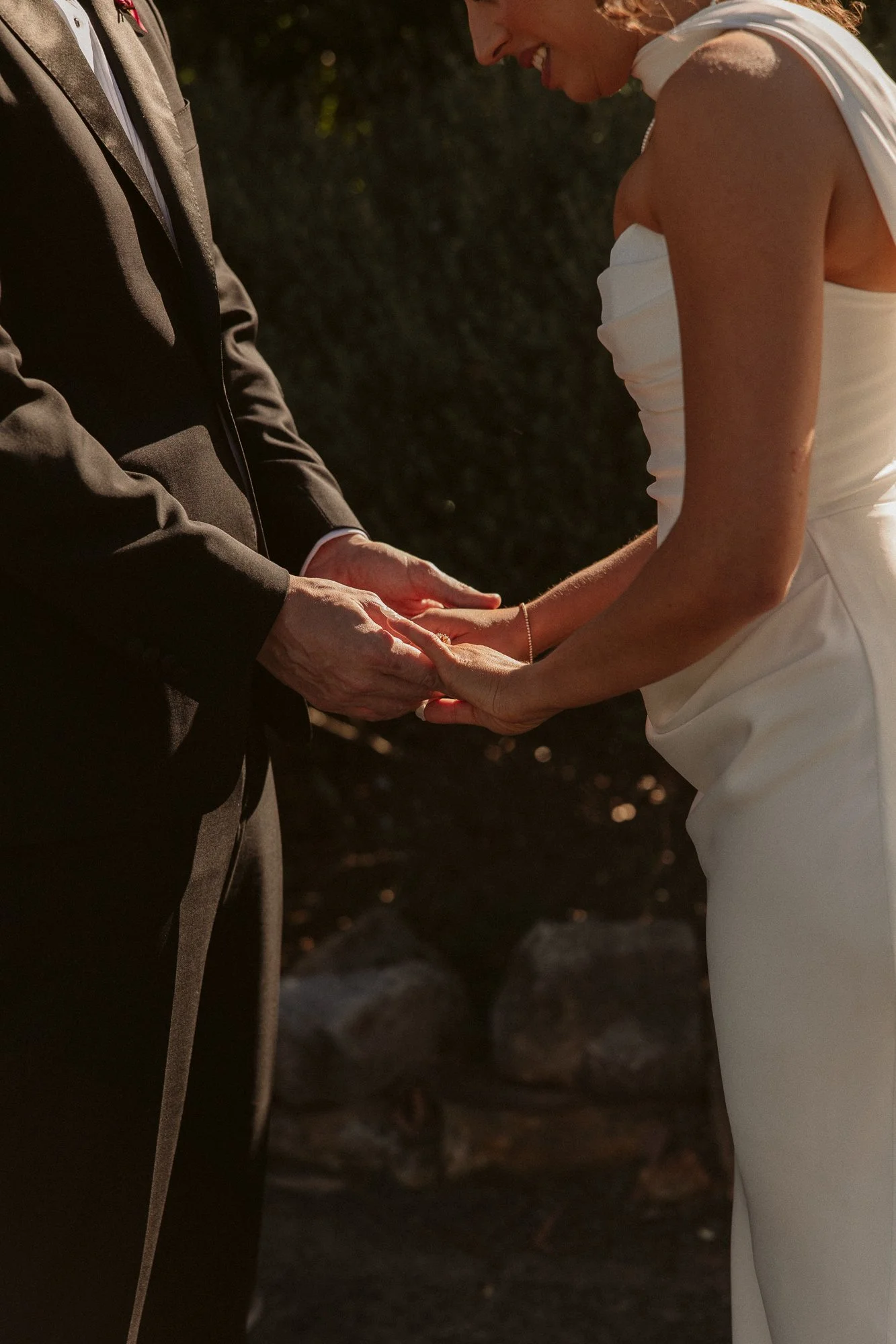 A bride and groom holding hands during their wedding ceremony, with the groom in a dark suit and the bride in a white dress, outdoors with sunlight and trees in the background.