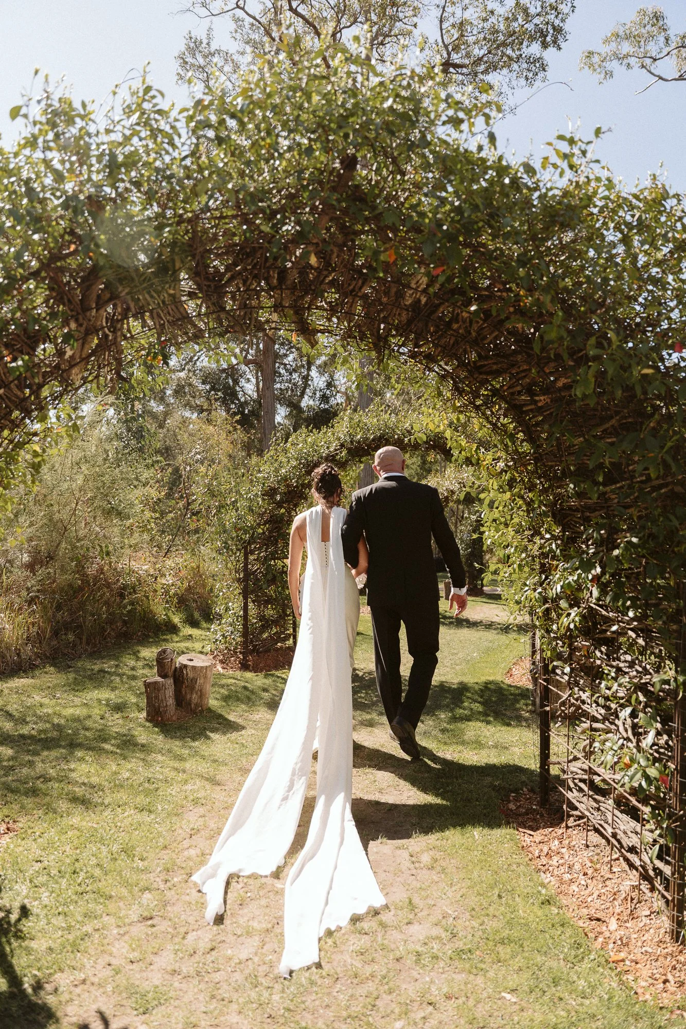Bride and groom walking through a garden archway on their wedding day, holding hands.