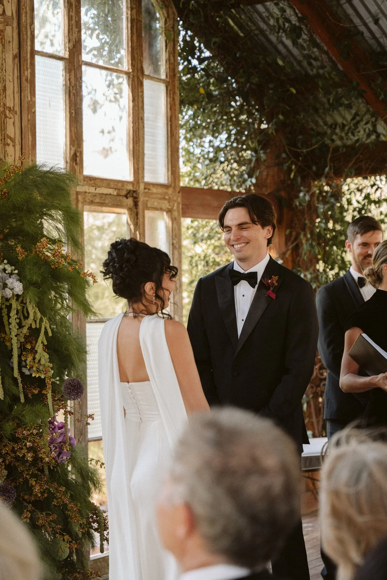 A bride and groom smiling at each other during their wedding ceremony indoors with large windows and greenery.