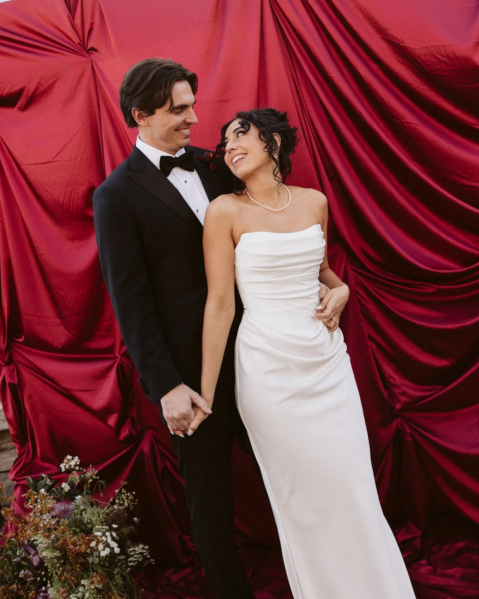A bride and groom holding hands and smiling at each other in front of a red draped fabric backdrop. The groom is wearing a black tuxedo with a bow tie, and the bride is in a white strapless wedding gown with pearl necklace and black curly hair.