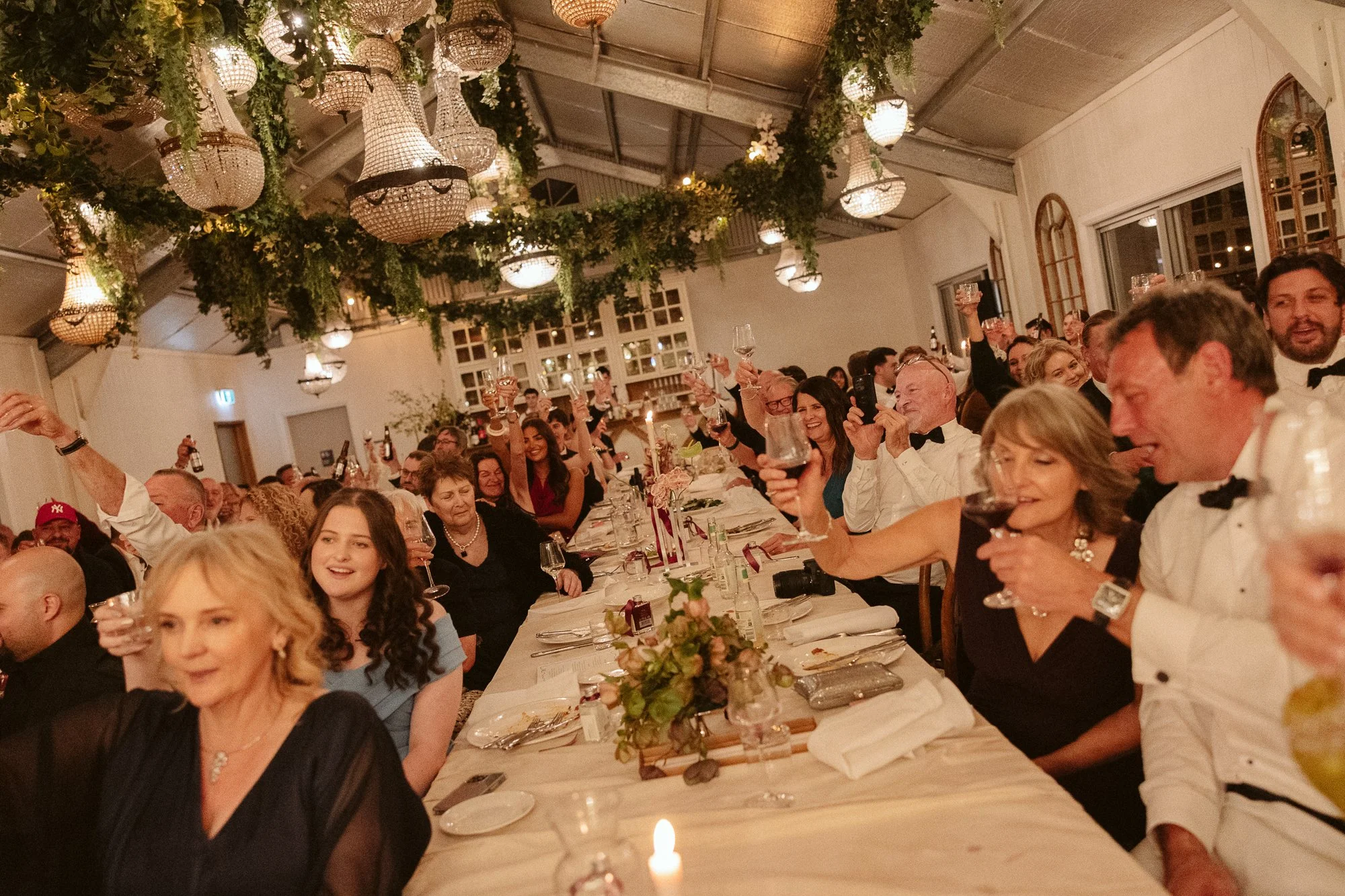 A group of people at a banquet table raising glasses in cheers at a formal celebration, under chandeliers and greenery decorations.