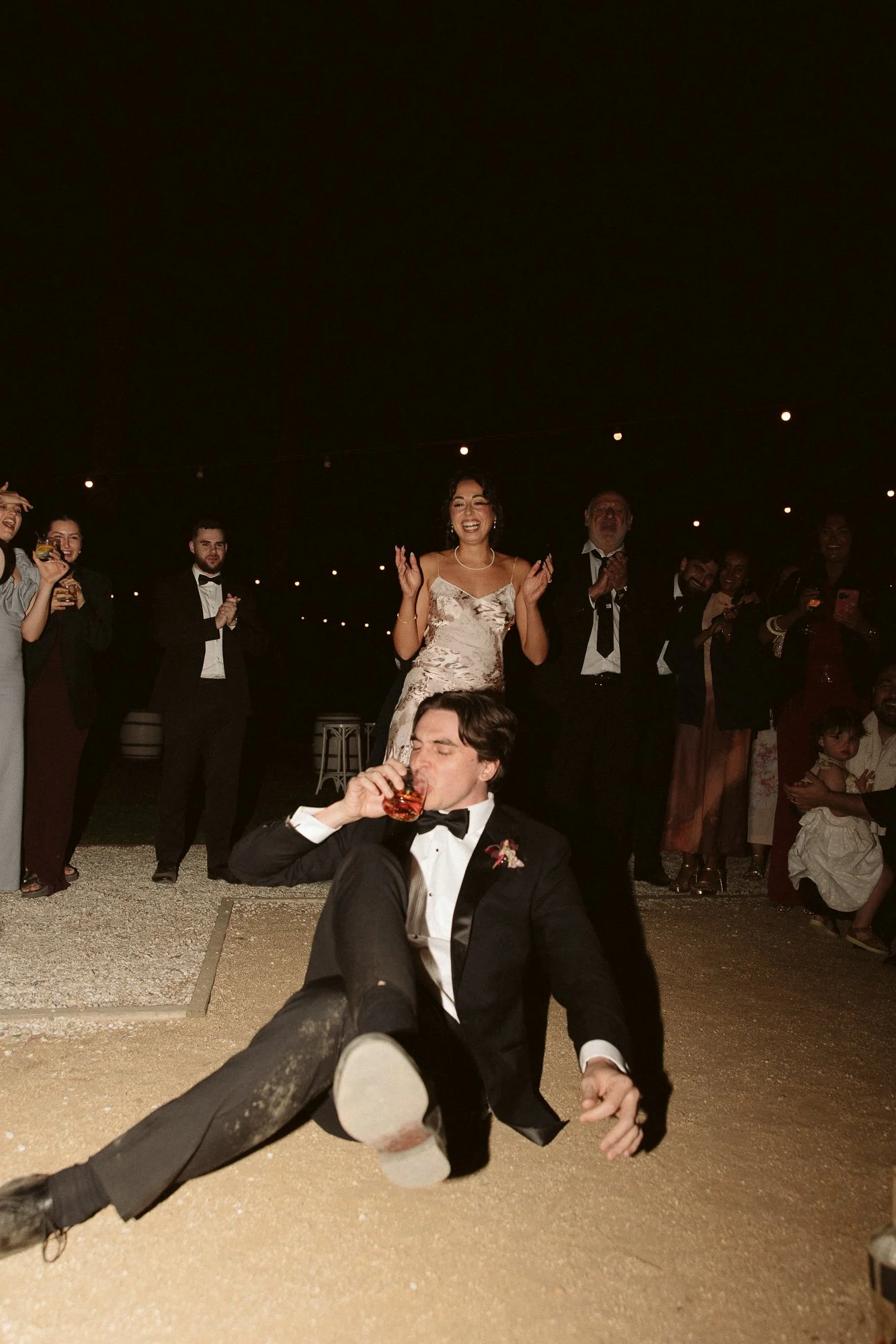 A man in a tuxedo is sitting on the ground drinking from a glass, with a woman in a wedding dress and guests in the background clapping and smiling at night.