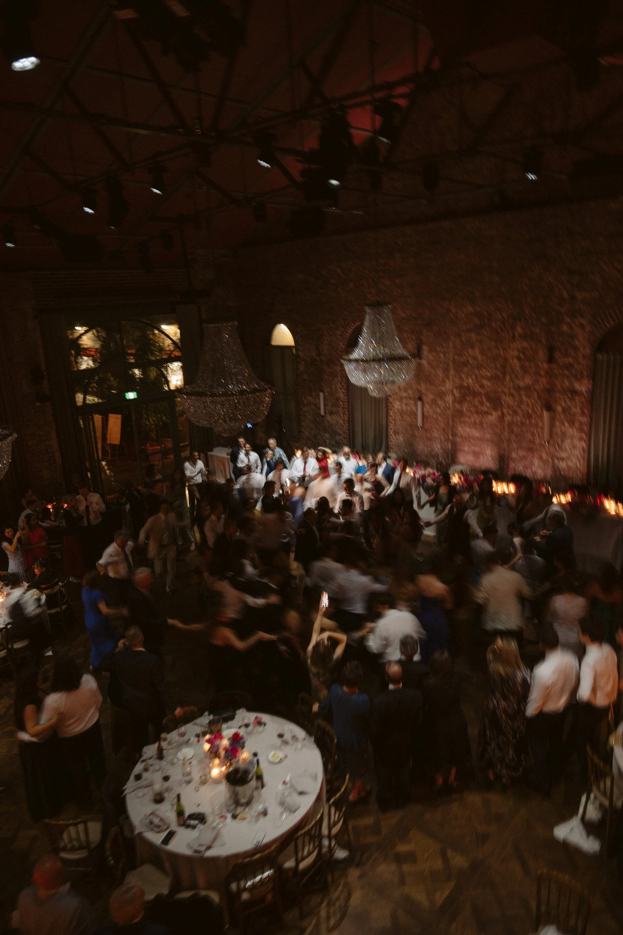 People dancing at a wedding reception in a dimly lit venue with brick walls, large chandeliers, and a decorated round table nearby.