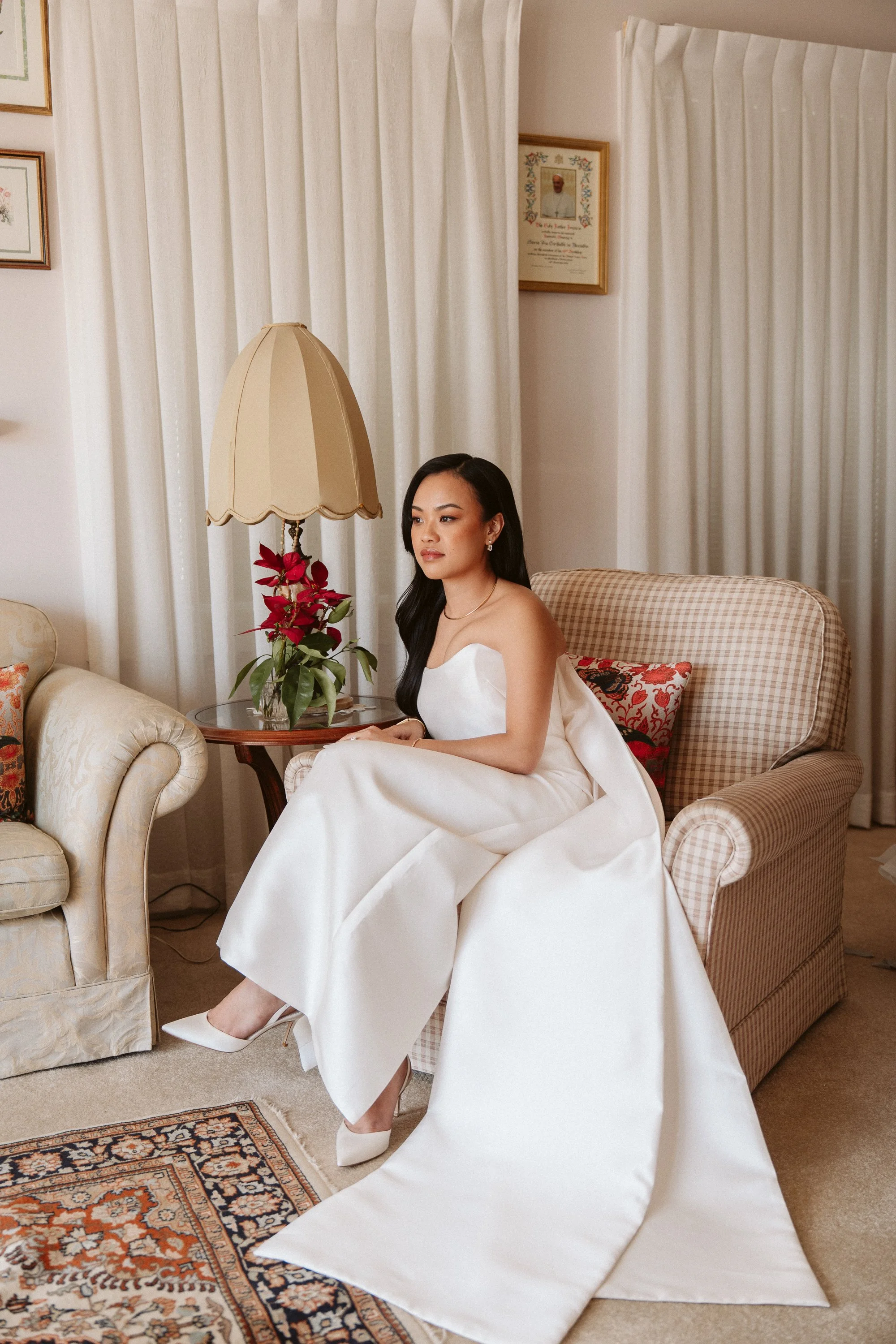 A woman in a white dress sitting on a plaid armchair next to a side table with a lamp and a plant, in a living room with framed pictures and curtains.