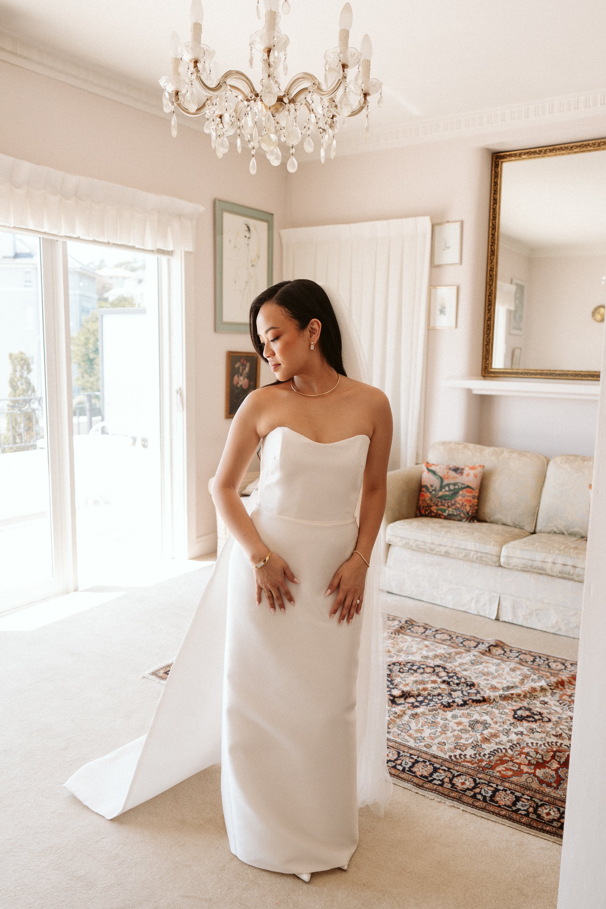 A bride in a strapless white wedding gown stands in a bright, elegant living room with a chandelier, a large mirror, and a patterned rug.